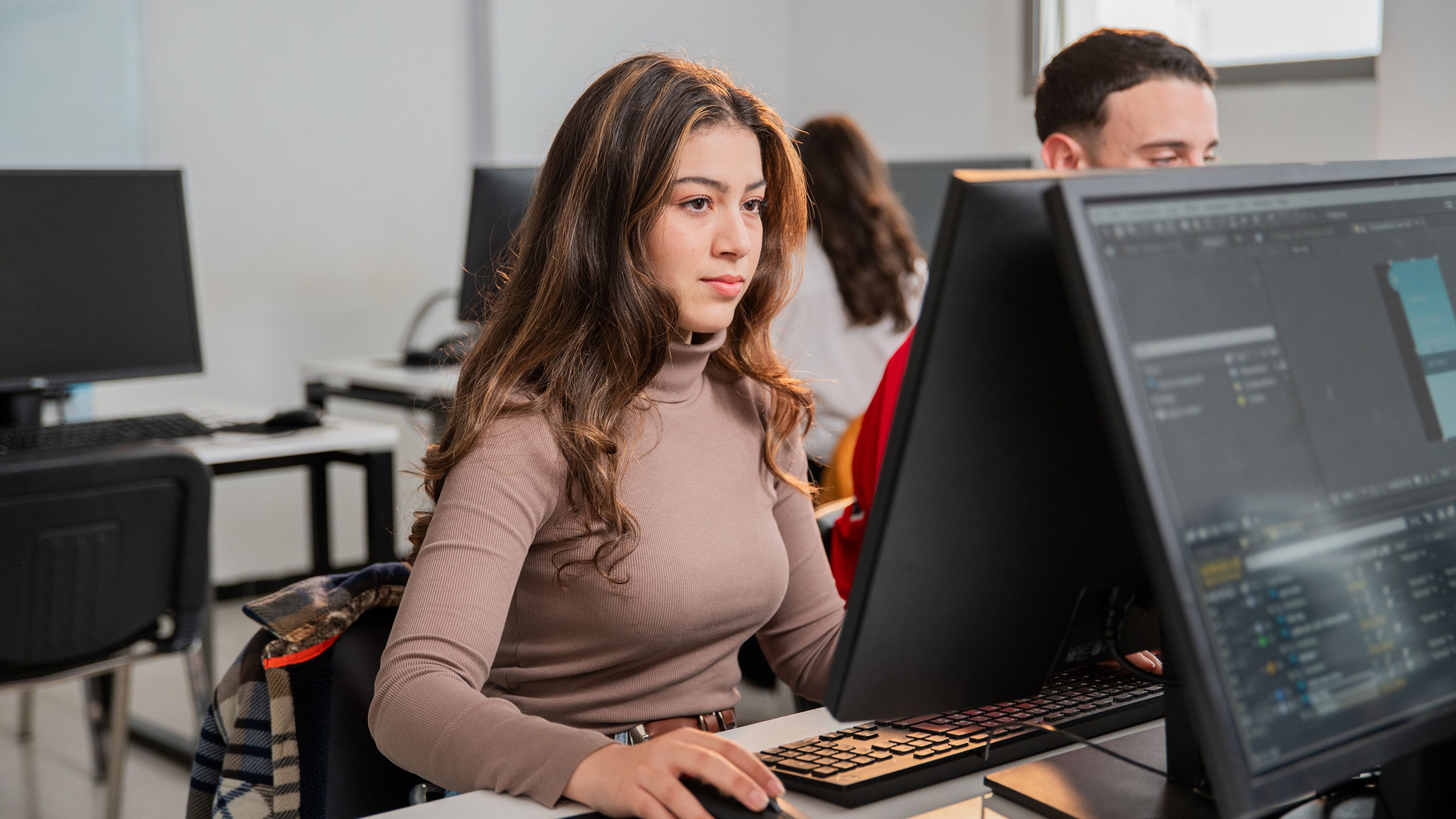 Une jeune femme concentrée aux longs cheveux travaille sur un ordinateur dans un bureau, avec des collègues en arrière-plan.