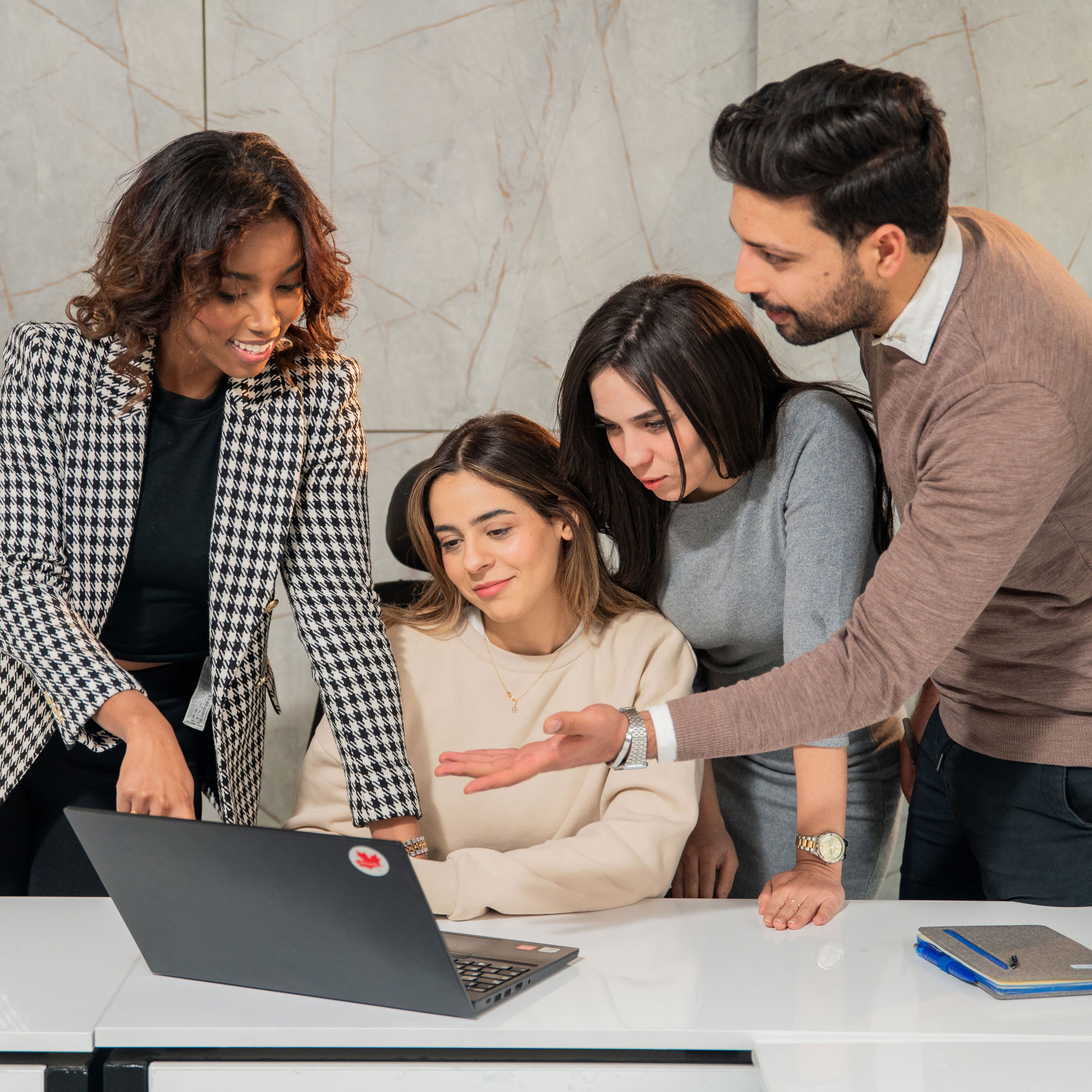 Four professionals gathered around a laptop, actively engaged in a discussion, with positive expressions and a modern office background.