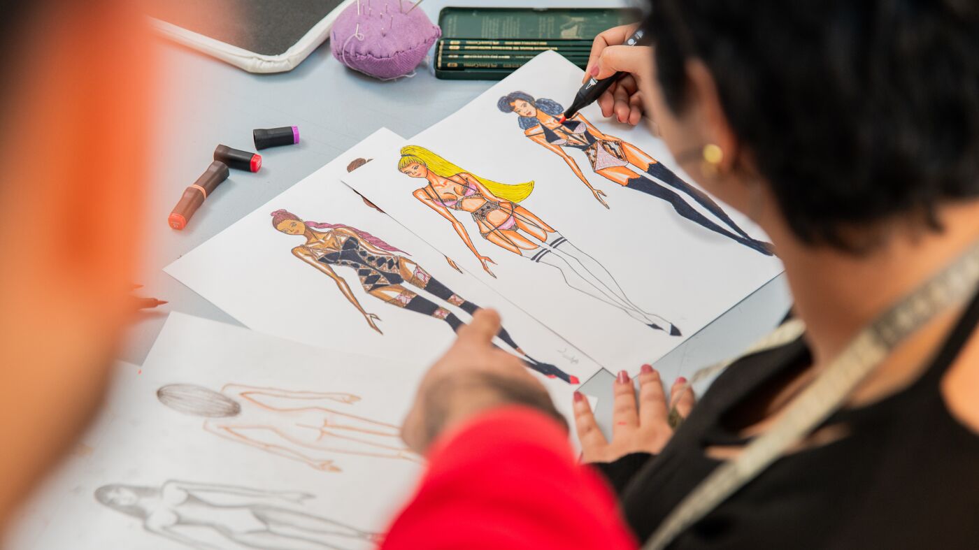 A person crafts detailed fashion illustrations with markers on a desk strewn with drawing tools.