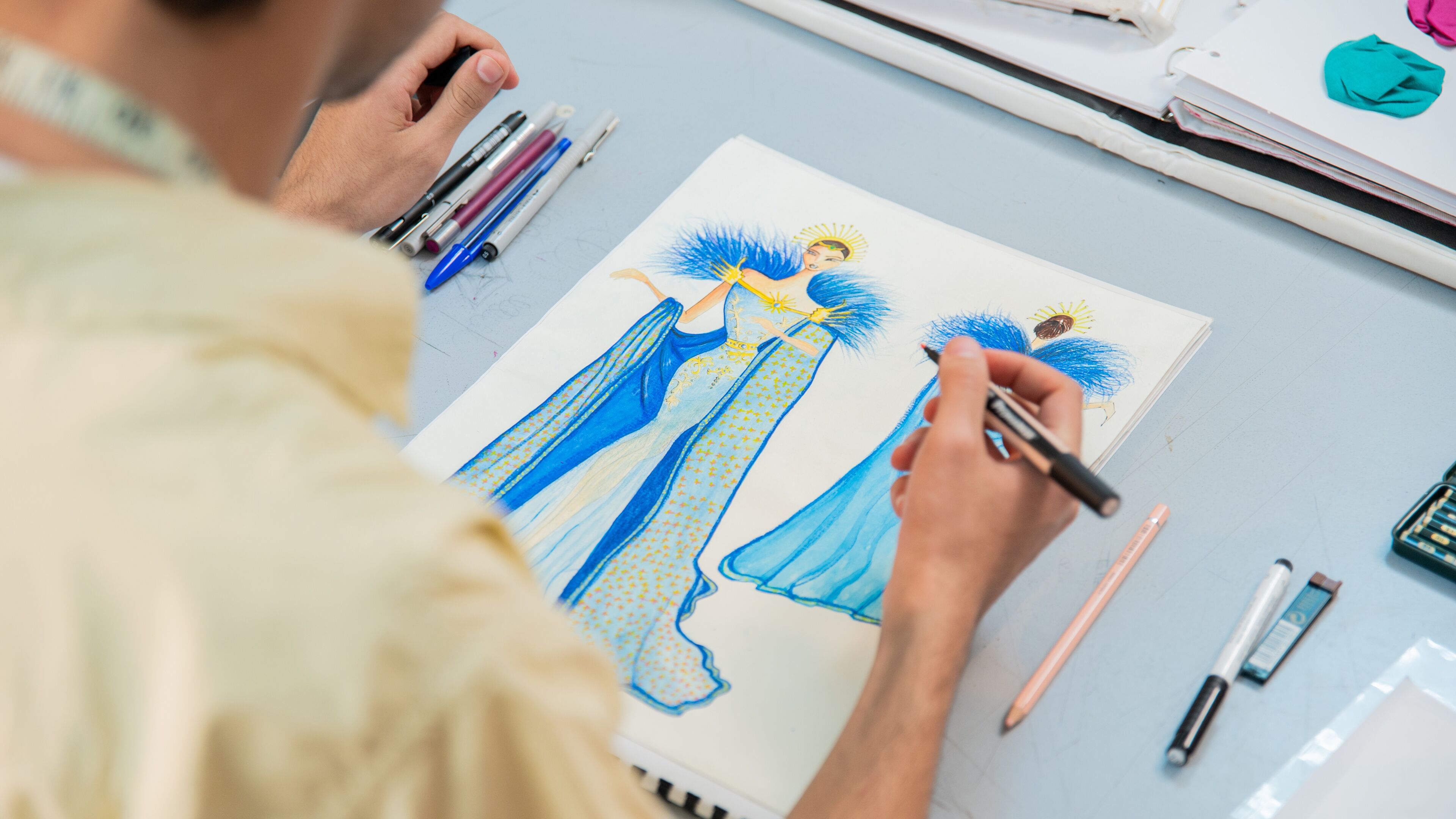 Un(e) designer dessine une illustration détaillée d'un costume bleu et doré avec des plumes et une coiffe en halo.
