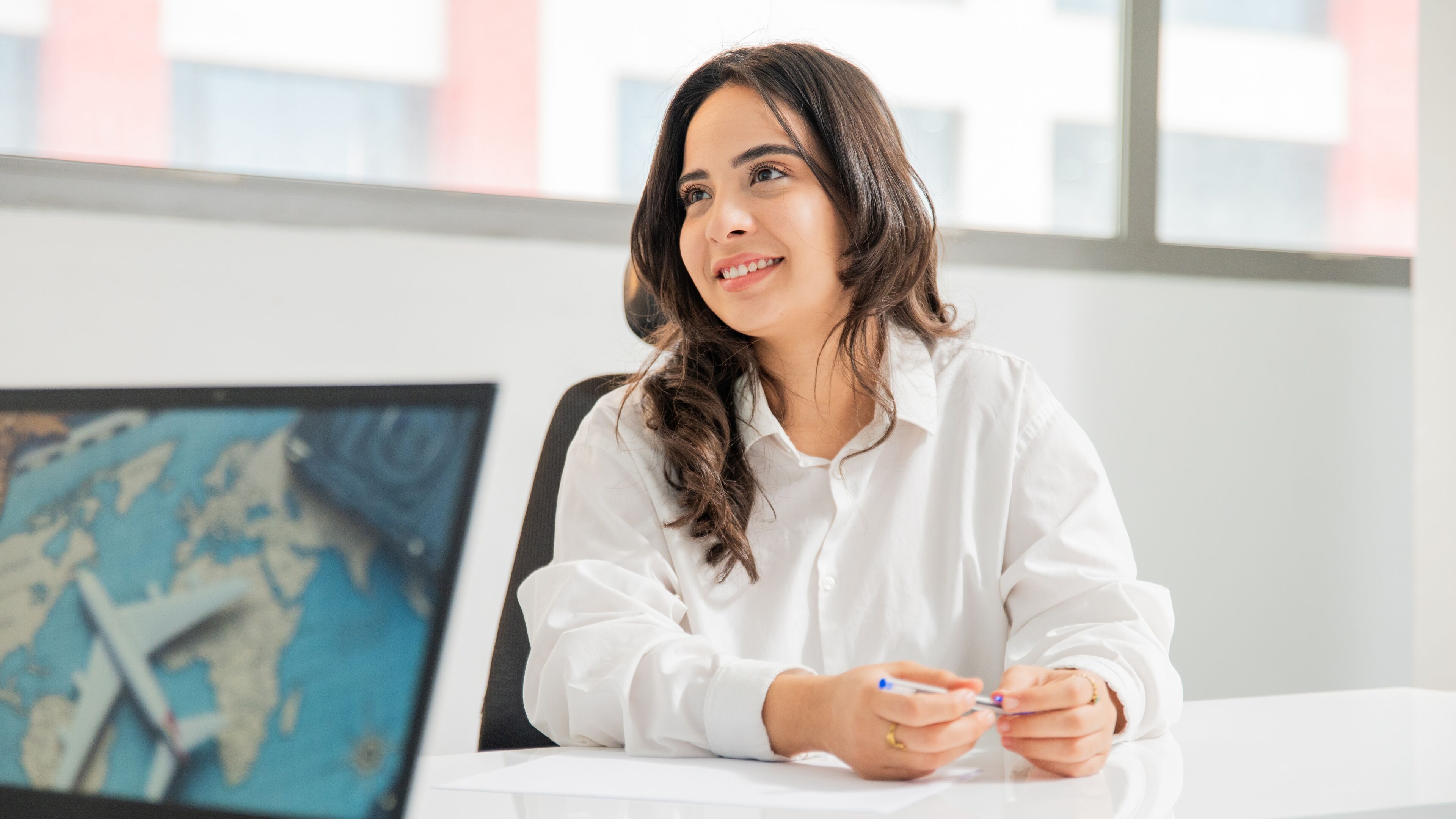Une femme souriante aux longs cheveux bruns, vêtue d'une blouse blanche, assise à un bureau dans un bureau lumineux, avec une carte du monde sur son écran d'ordinateur, transmettant un sentiment d'engagement commercial mondial.
