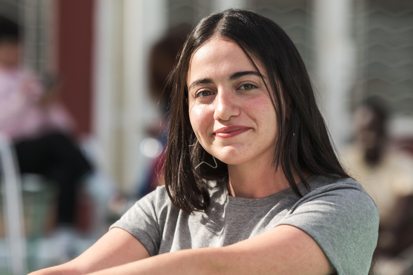 A young woman with straight dark hair and a gray t-shirt smiles warmly while sitting outdoors. The blurred background with natural light highlights her relaxed and cheerful demeanor in a casual setting.