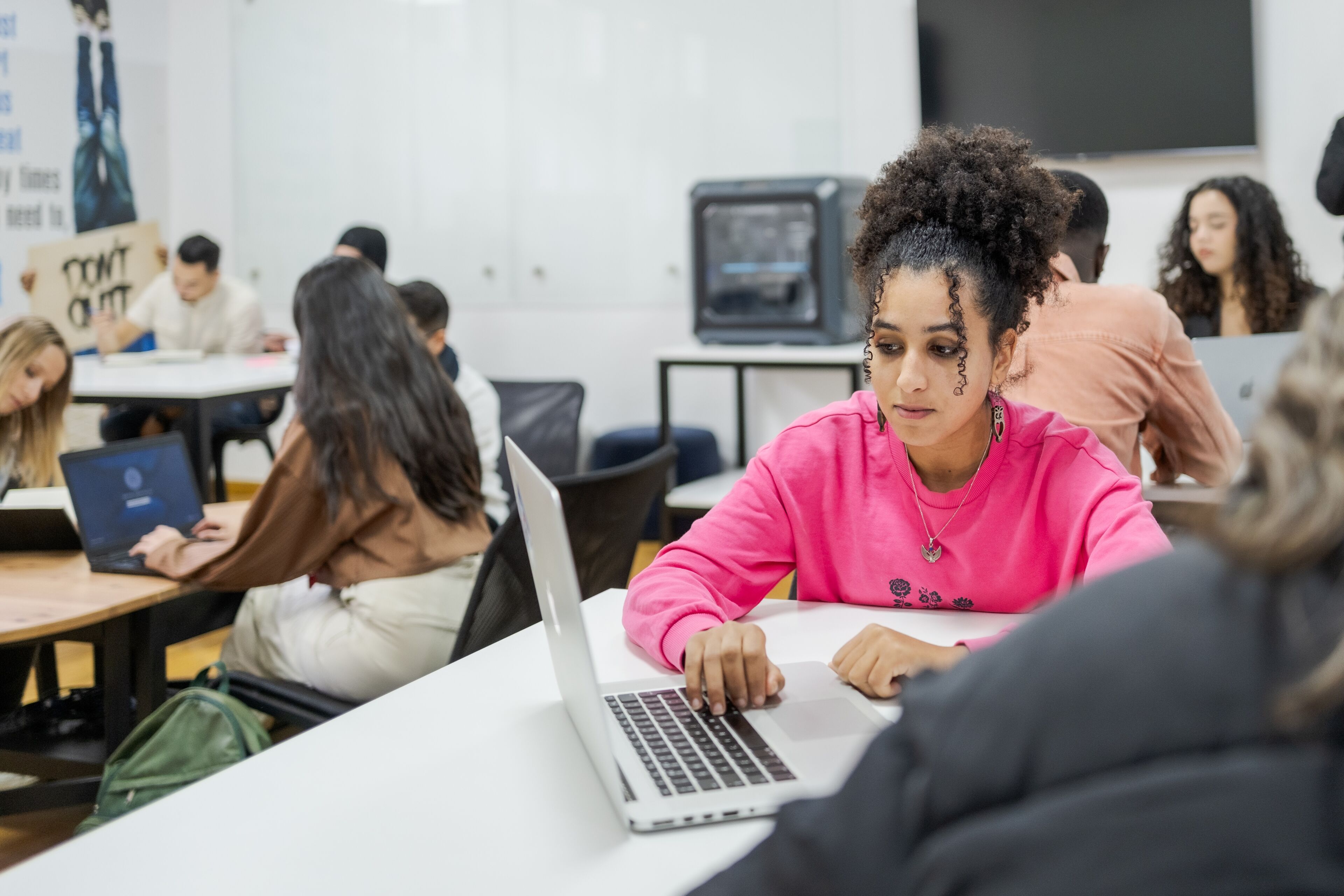 A young woman in a pink sweatshirt works on her laptop in a classroom filled with other focused students.