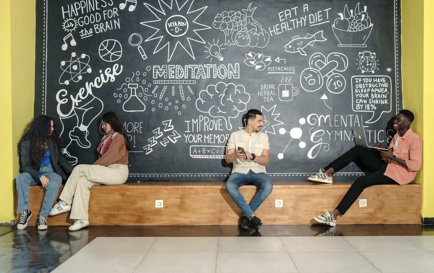 Four young adults casually sit and chat in front of a detailed chalkboard featuring illustrations and tips about health, exercise, and mental well-being. The vibrant background adds a creative and inspiring touch to the relaxed group interaction.