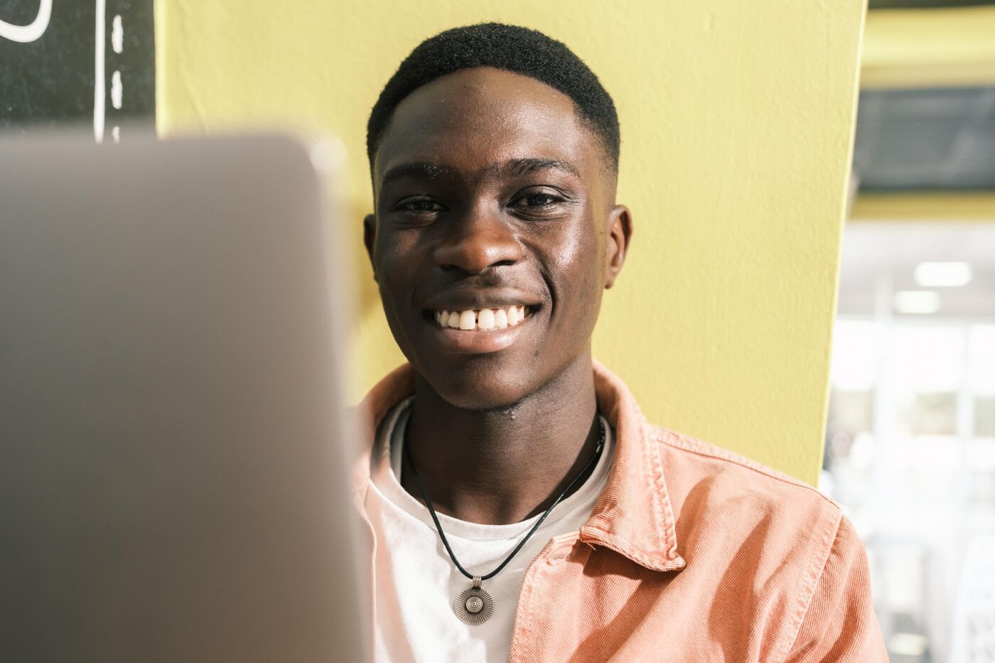 In the image, a young man is sitting in front of a laptop, smiling and looking at the camera.