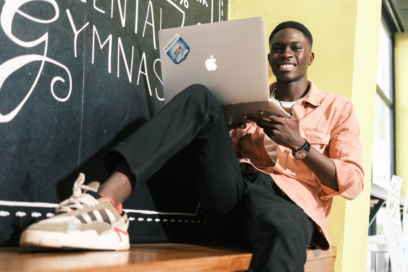 A young man wearing a peach-colored shirt and black jeans smiles while working on a laptop. He is seated casually indoors against a chalkboard-style wall, creating a relaxed and productive atmosphere.