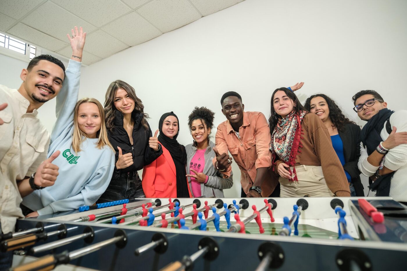  A cheerful and diverse group of friends posing around a foosball table, giving thumbs up and smiling. The casual setting in a bright room highlights their camaraderie and enjoyment of a fun social activity.