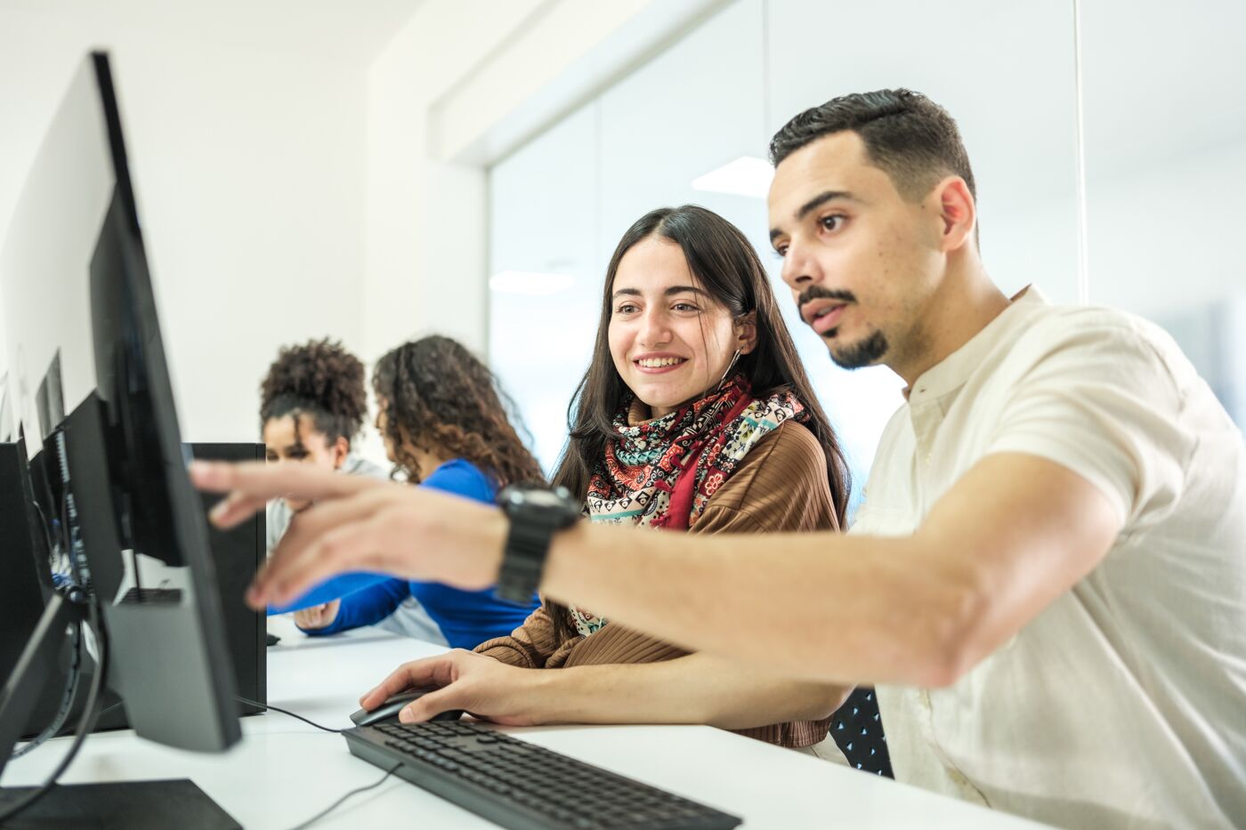 Two students engaging with a computer task, working together in a computer lab with peers focused in the background.