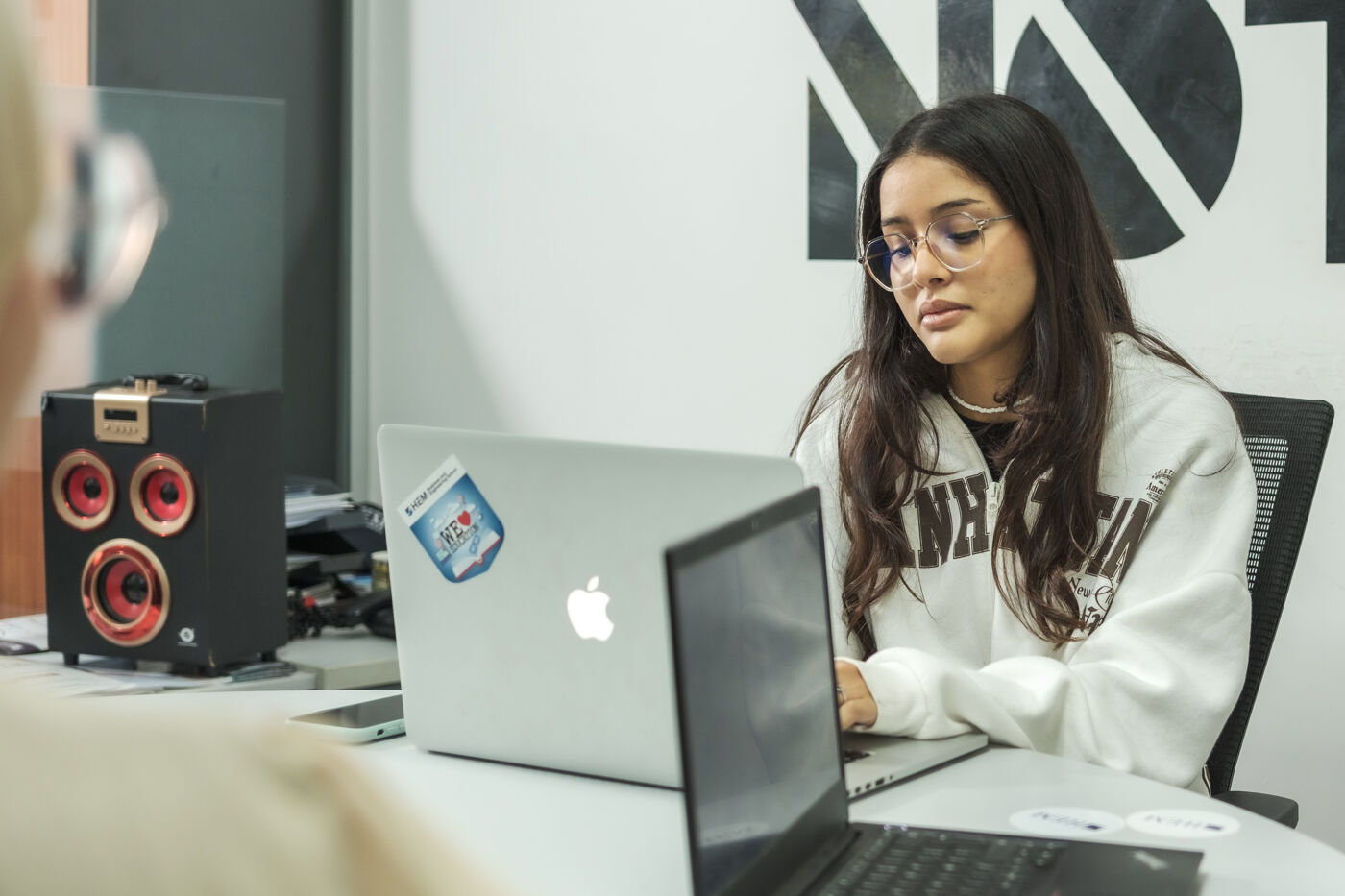 A focused woman with glasses works on a MacBook in an office setting, a speaker nearby and decor suggesting a creative workspace.