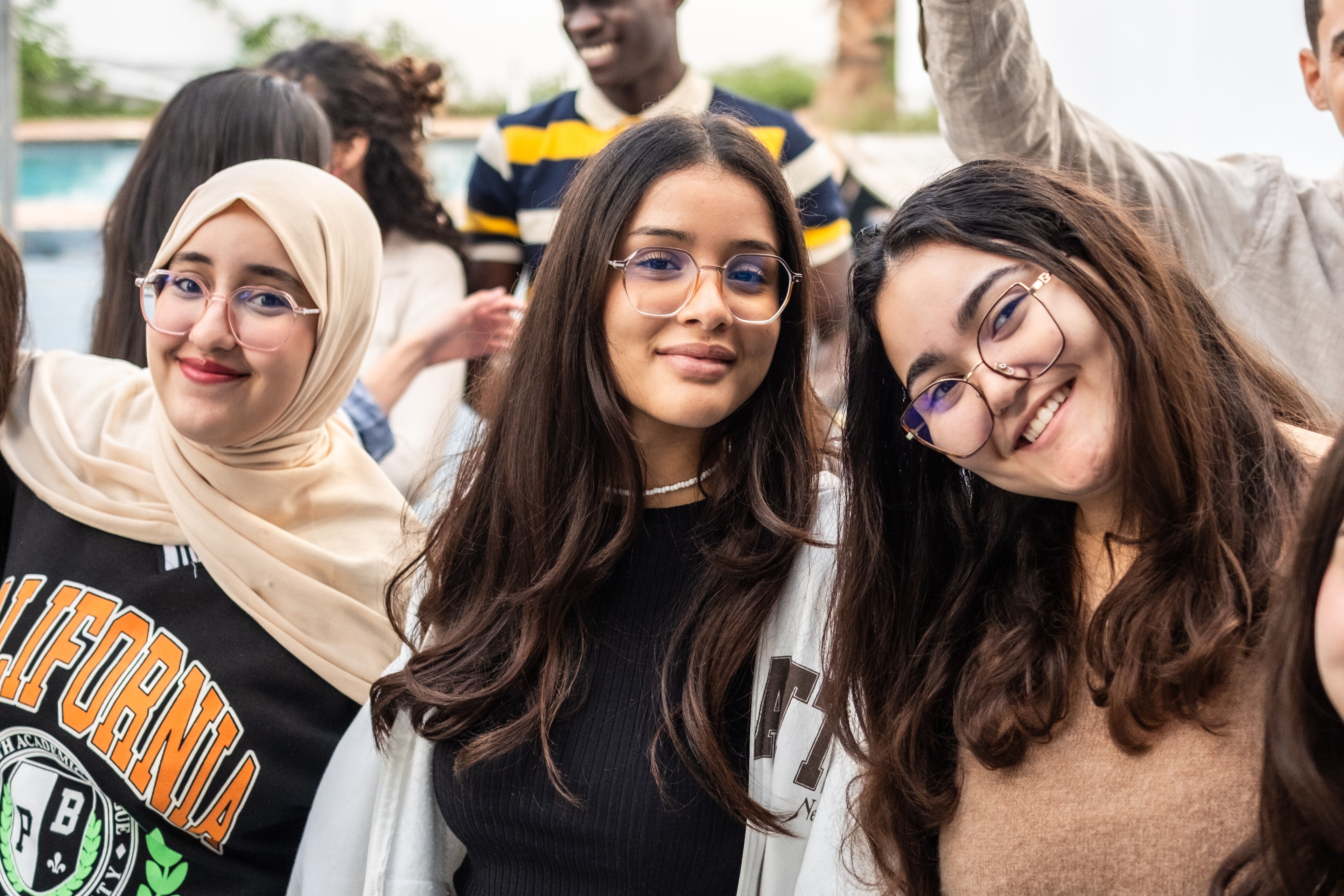 Three smiling young women at an outdoor gathering, with one wearing a hijab.