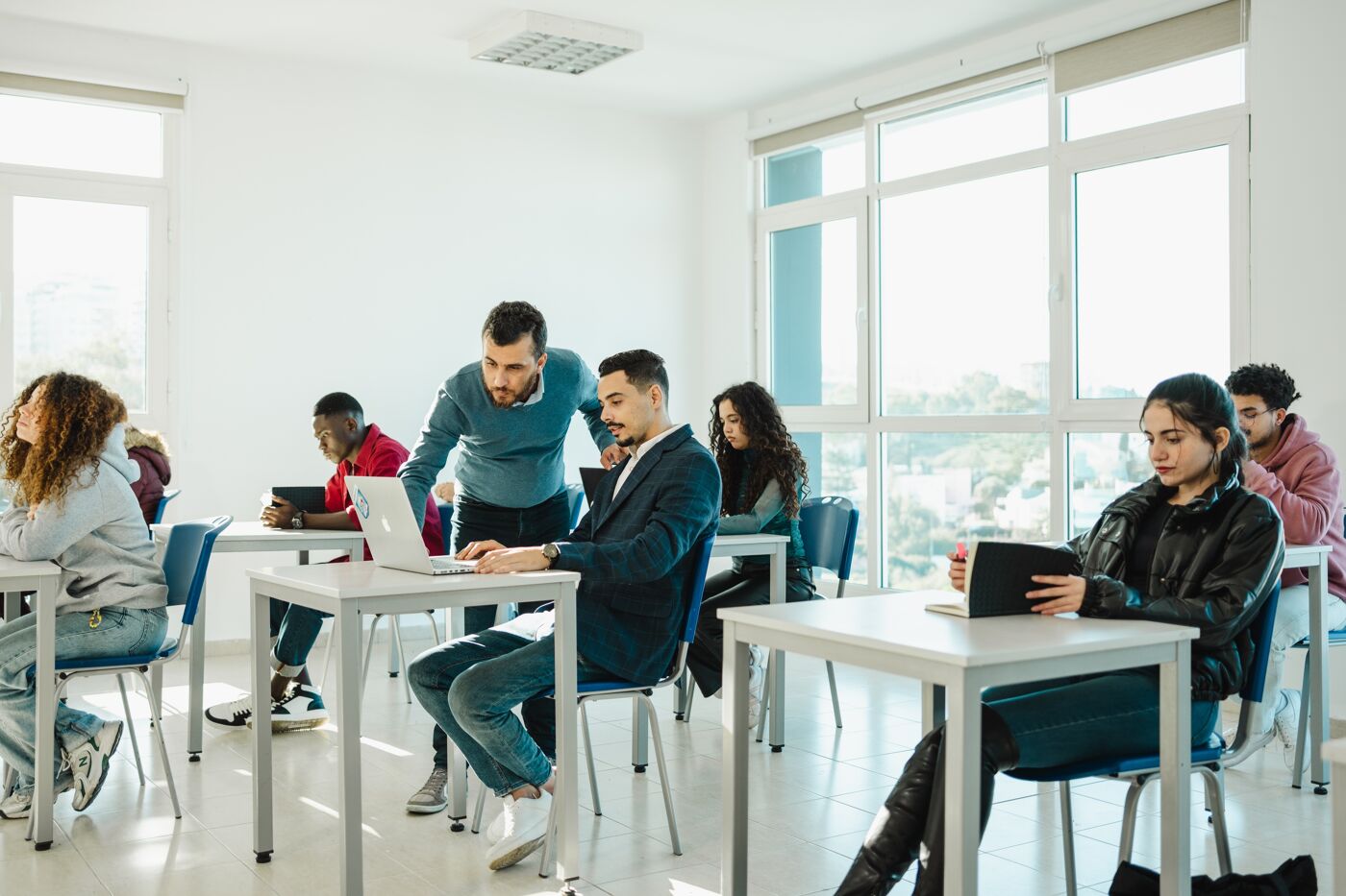 Diverse group of students studying with laptops and notebooks in a bright classroom setting, one of them assisting another.