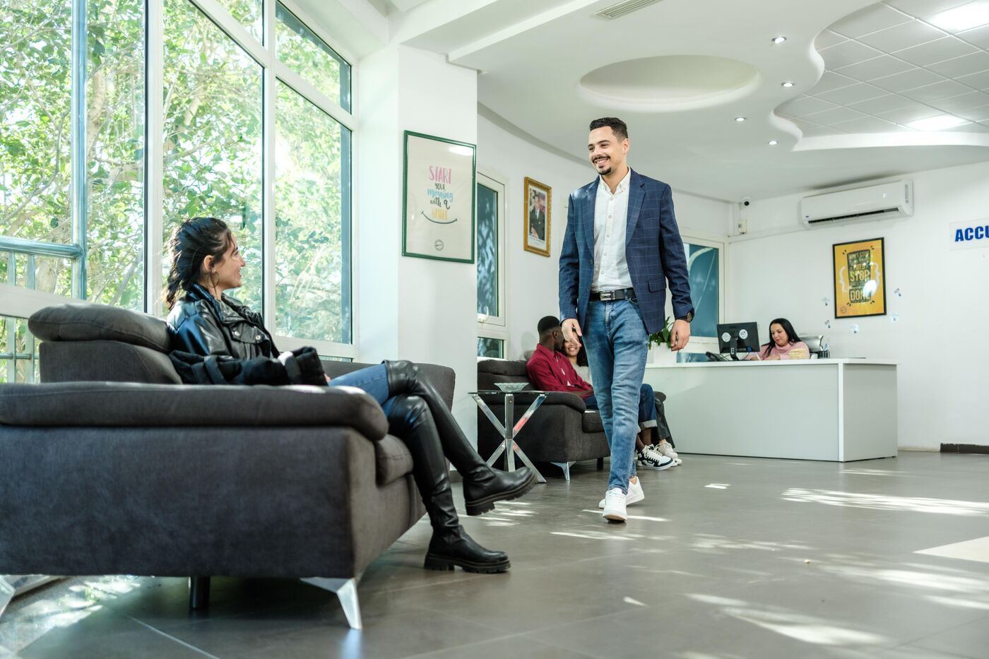 A well-dressed man walks confidently through a bright office lobby, exchanging smiles with a woman seated on a couch. Other individuals sit comfortably in the waiting area, while a receptionist works in the background, creating a welcoming and professional setting.