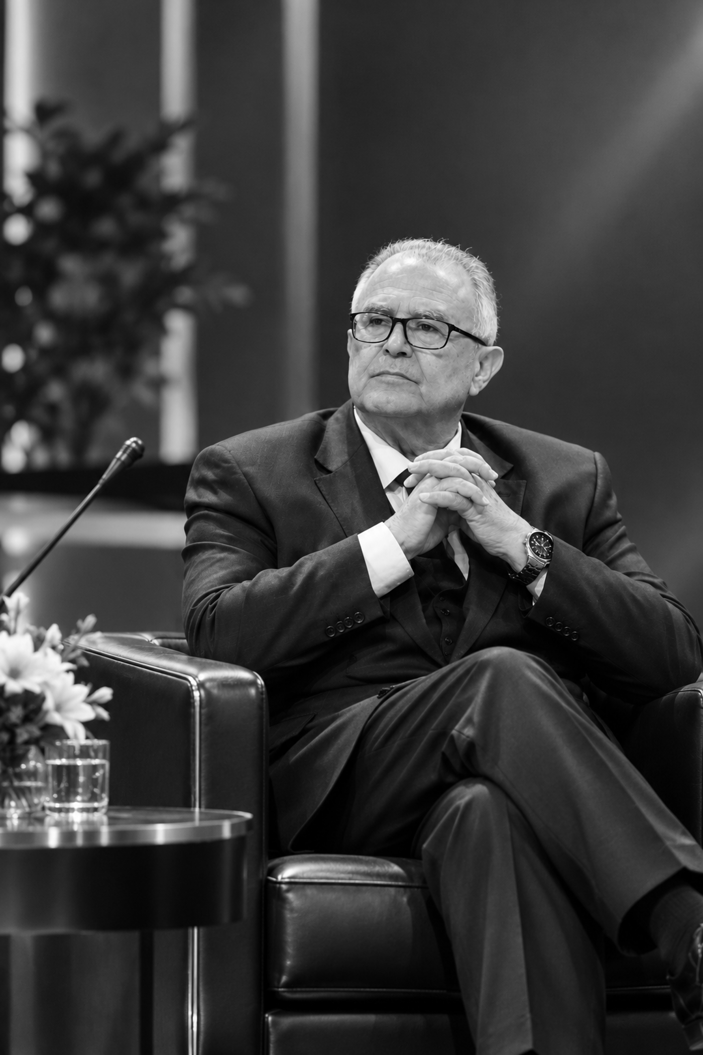 A distinguished older gentleman in a suit and tie sits attentively, with his hands clasped and gaze directed thoughtfully off-camera, suggesting a moment of contemplation during a formal event.