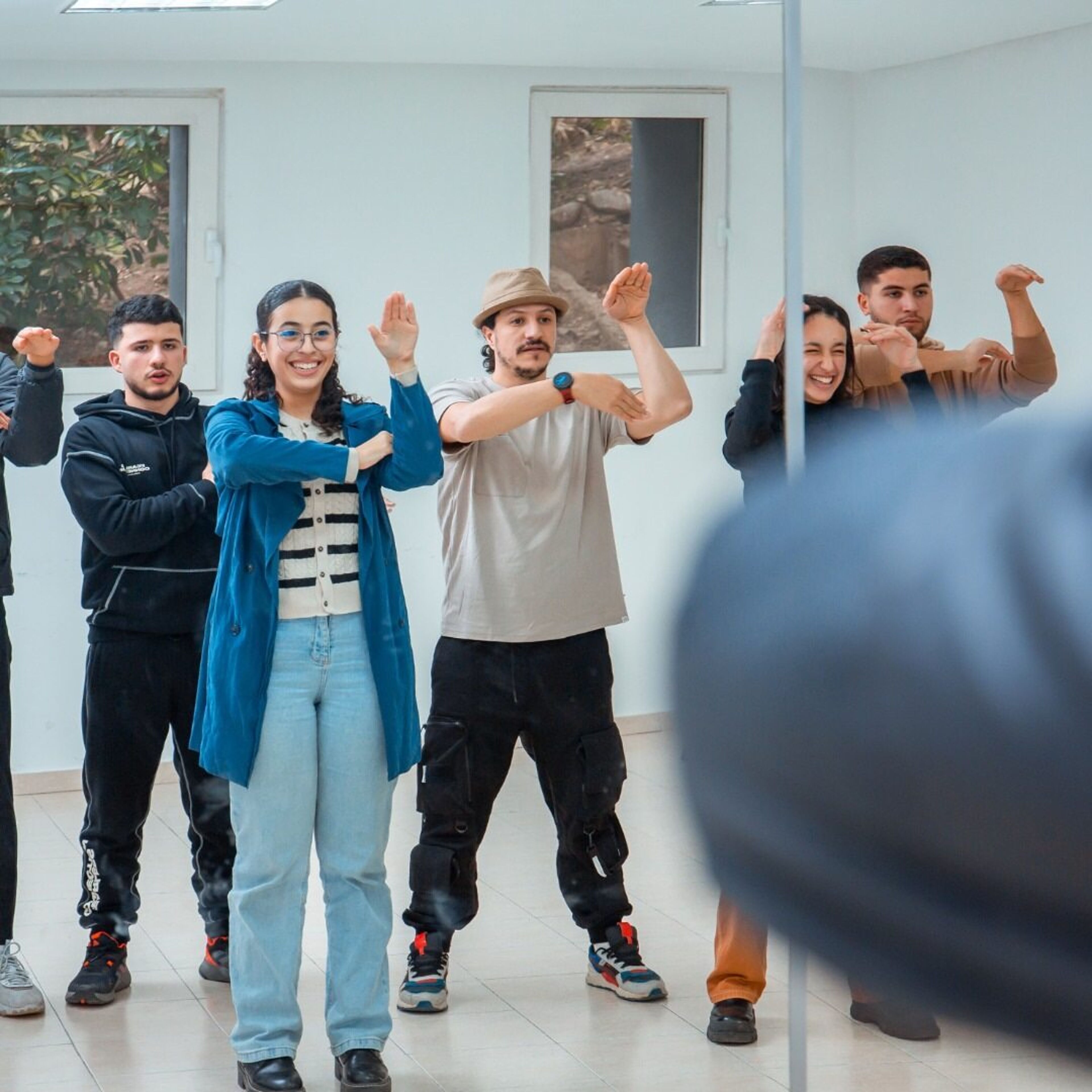 A group of young adults participates in a dance class, mirroring each other's arm movements in a bright studio with large windows. The participants appear engaged and cheerful, with one instructor leading the session. The casual attire suggests a relaxed, friendly atmosphere.