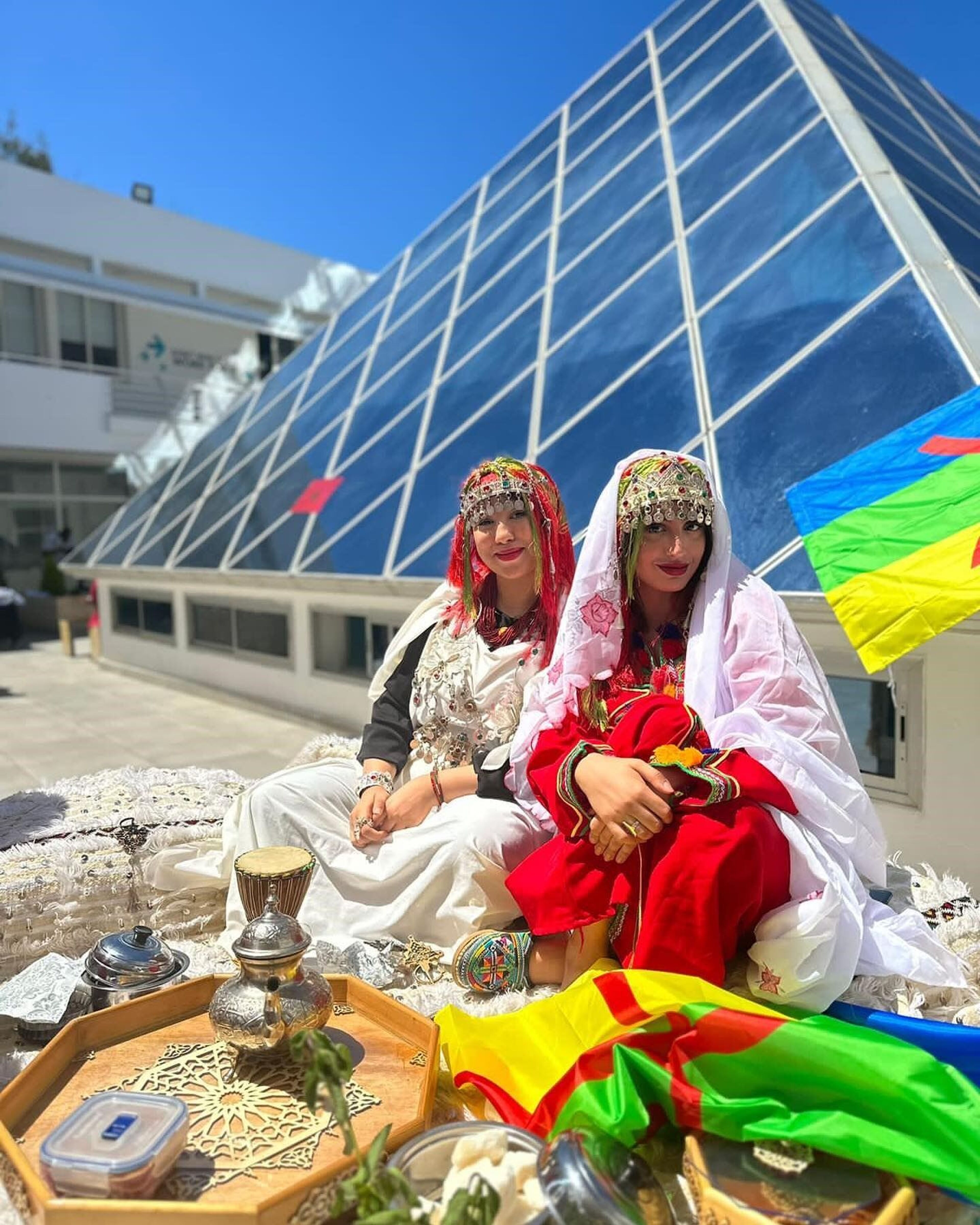 Two women dressed in traditional Berber clothing sit beside a glass pyramid structure, showcasing vibrant attire and intricate jewelry. The scene includes cultural items on a tray and a colorful Berber flag, highlighting a celebration of heritage under a clear blue sky.