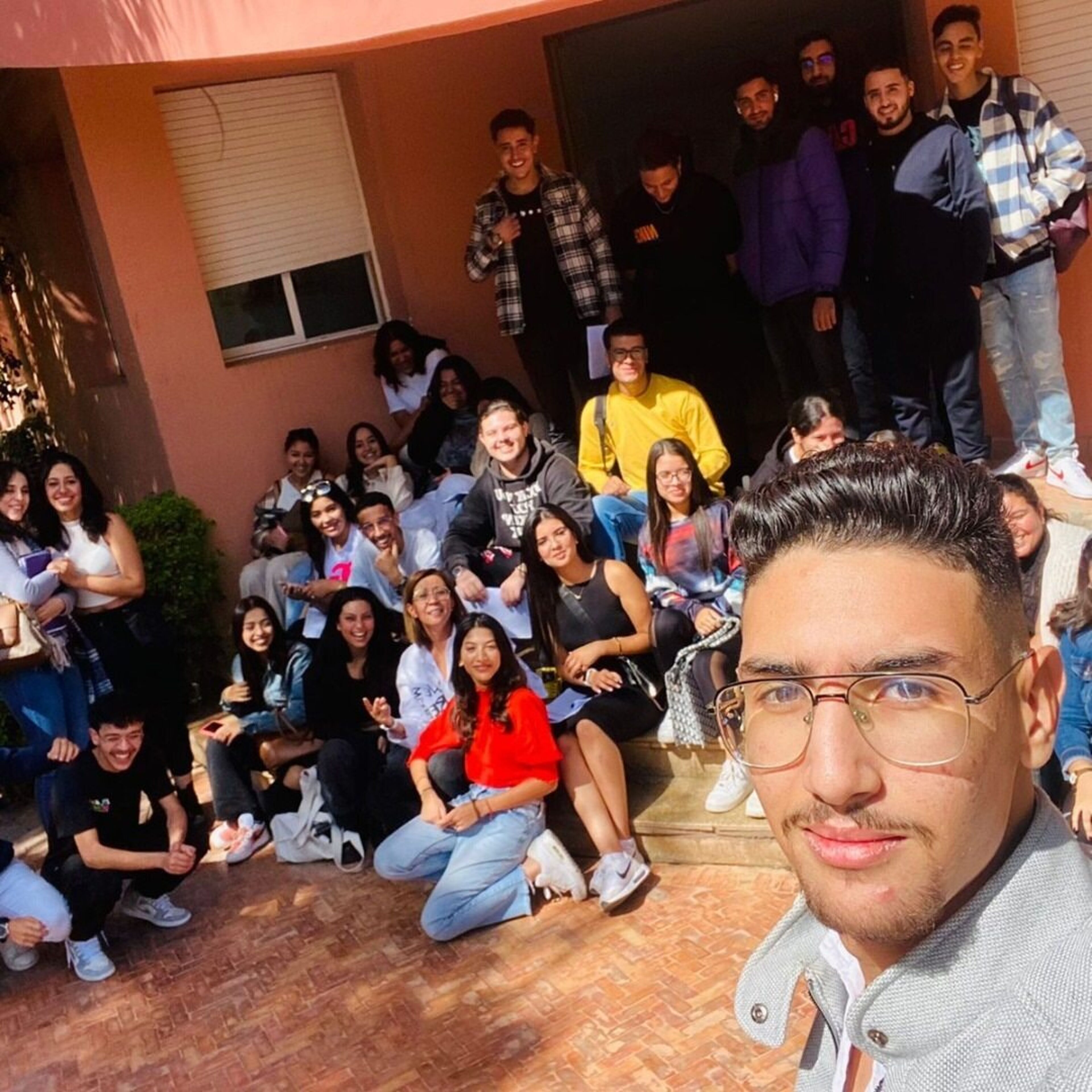 A large group of young adults gathers outside, smiling and posing for a group photo in the sun. They are casually dressed, sitting and standing around a building entrance. The atmosphere is cheerful and relaxed, suggesting a social gathering or informal meetup.