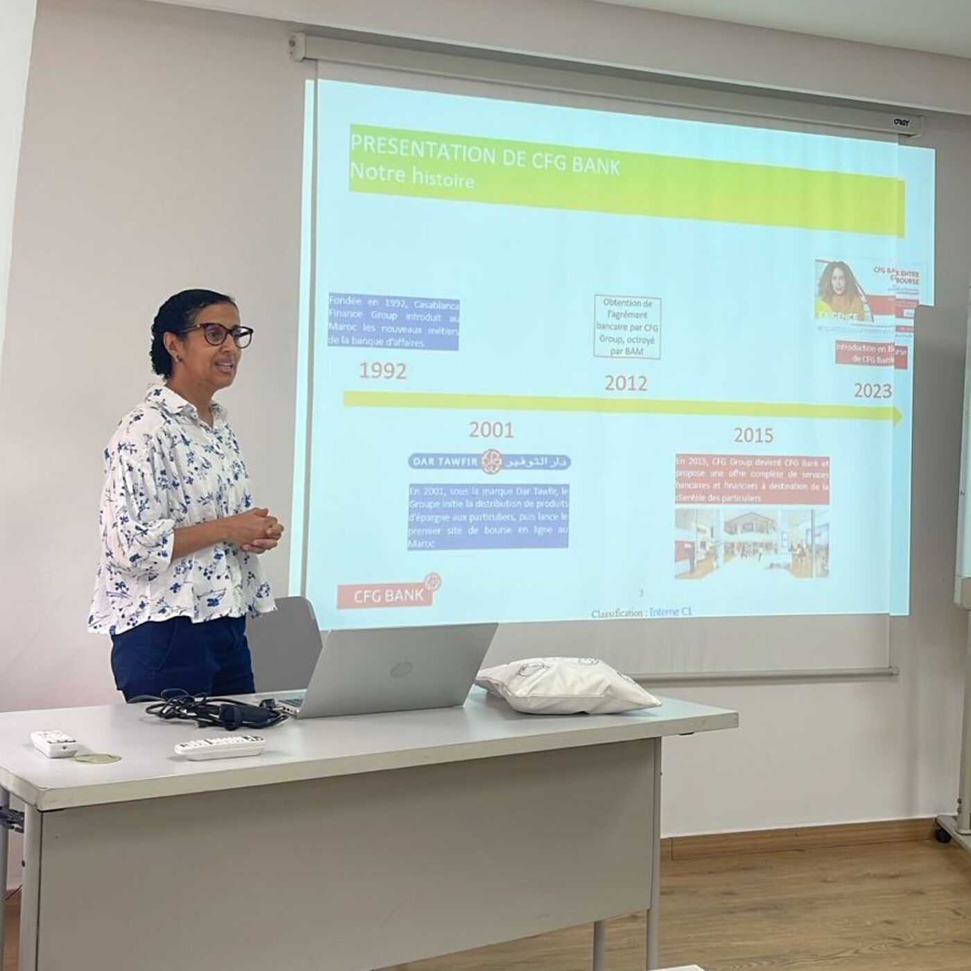 A woman is giving a presentation about CFG Bank's history, as shown on the projector screen behind her. The timeline includes milestones from 1992 to 2023, with various logos and text blurbs.