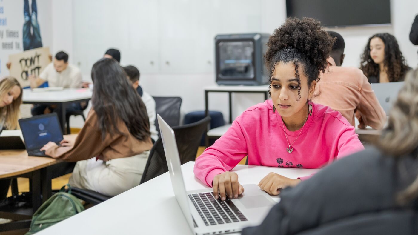 A young woman in a pink sweatshirt works on her laptop in a classroom filled with other focused students.