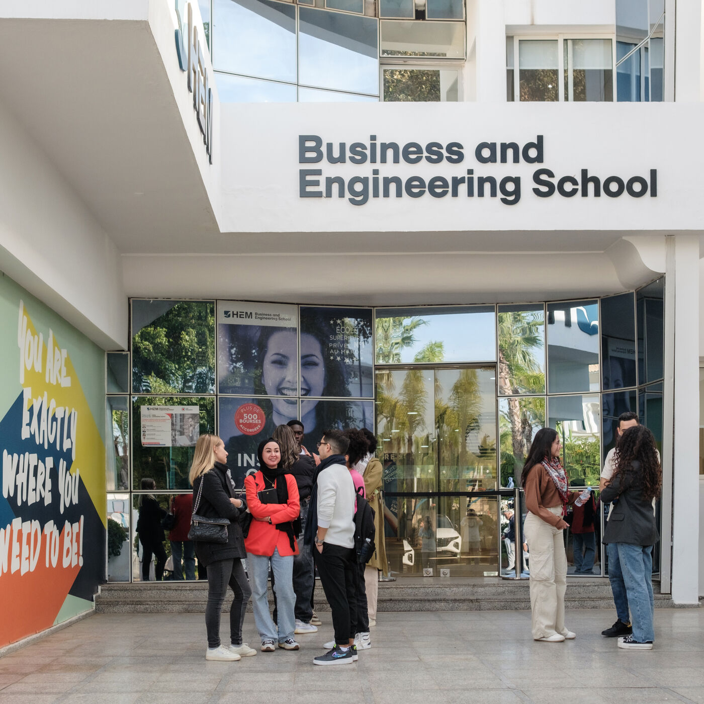 Students gather at the entrance of the Business and Engineering School, surrounded by modern architecture and a motivational banner.