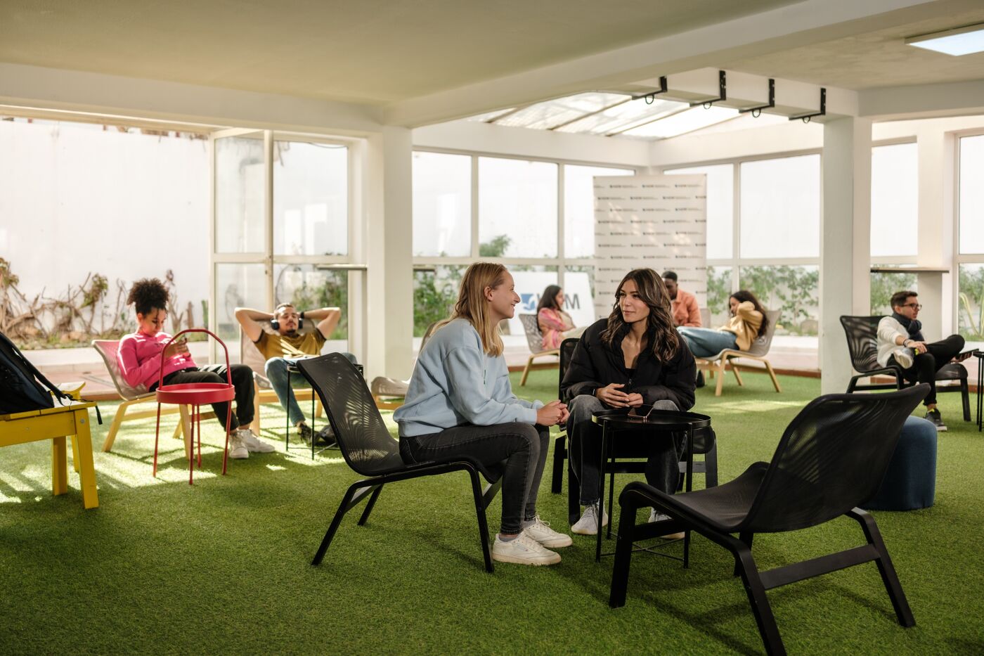 Individuals engaged in various activities in a bright office lounge with large windows and green flooring.