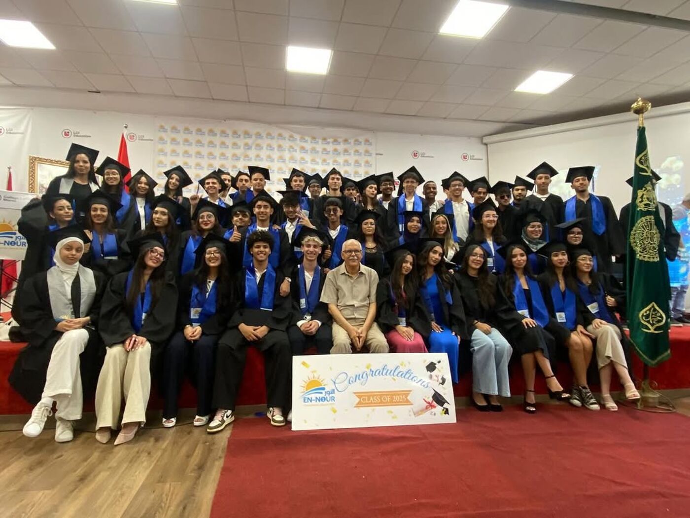 A group of graduates in caps and gowns pose for a photo with their professor, holding a congratulatory sign. They are standing on a red carpet in front of a white wall, with a flag on the right side.