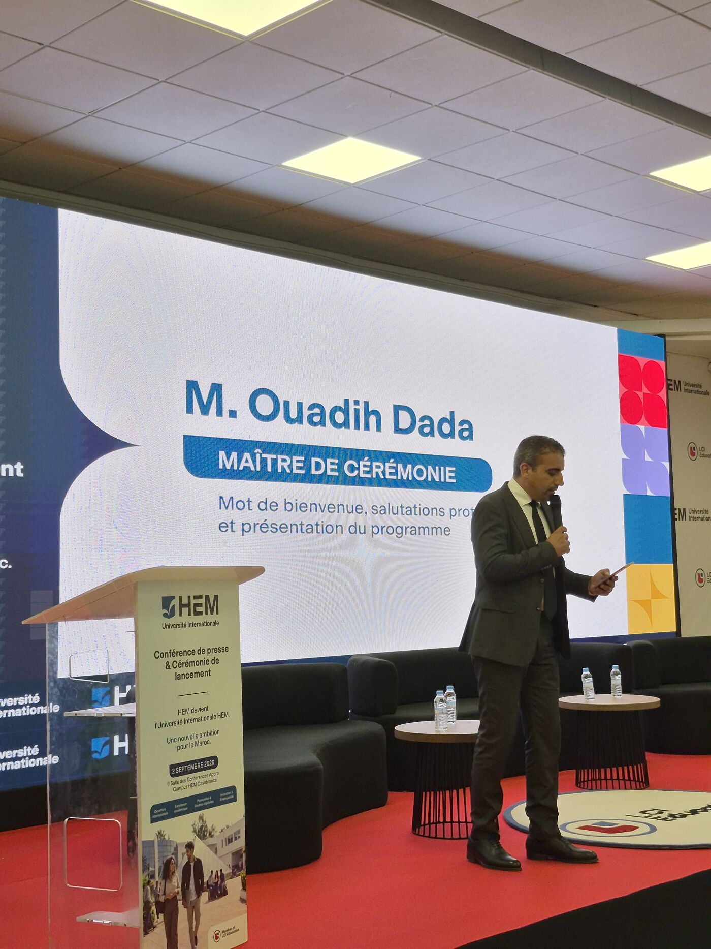 A man in a dark suit is speaking at a podium in front of a large screen that reads "M. Ouadih Dada, Master of Ceremonies".