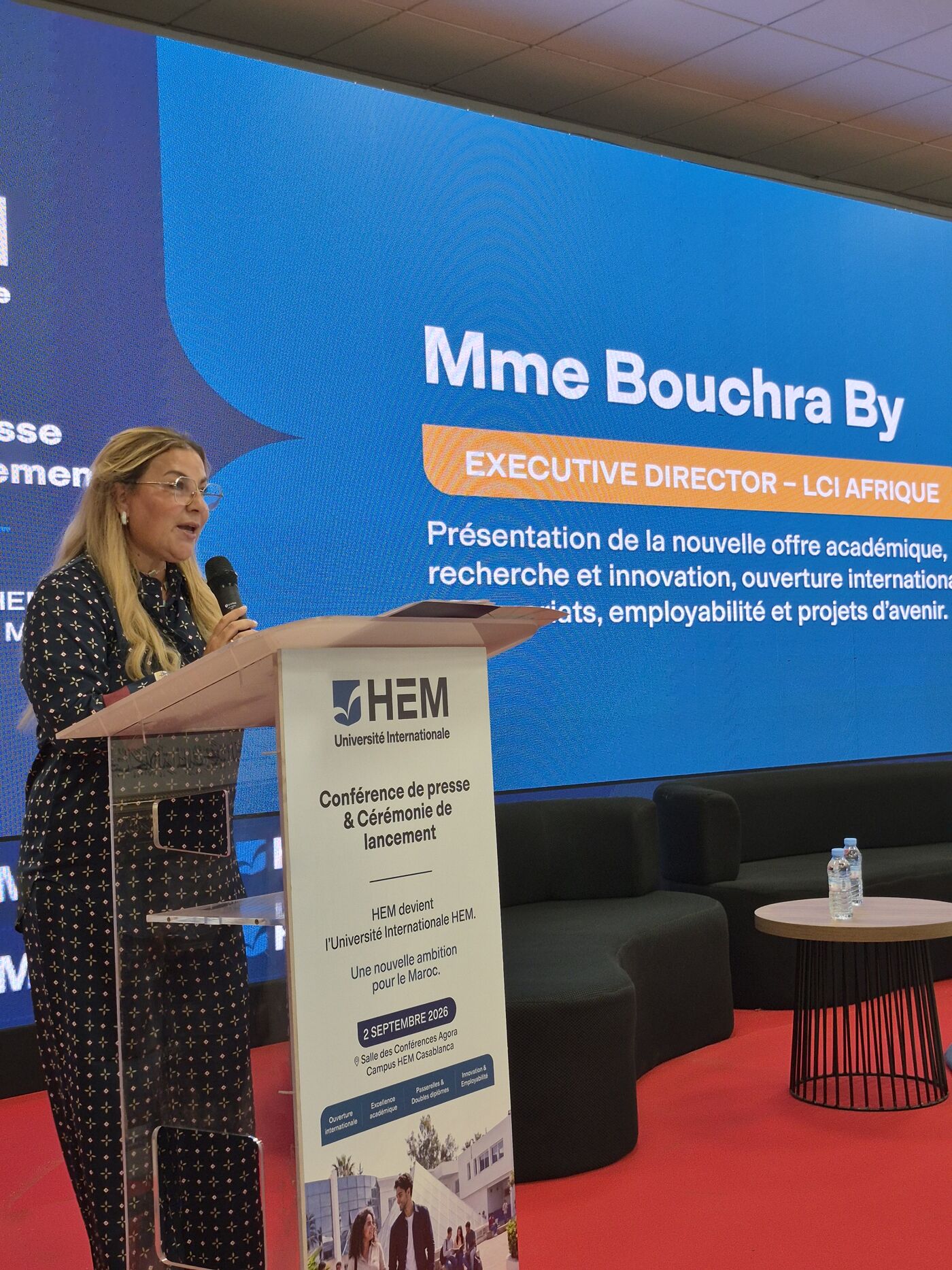 A woman speaks at a podium in front of a screen displaying "Mme Bouchra By EXECUTIVE DIRECTOR - LCI AFRIQUE". The conference is about a new academic offering.