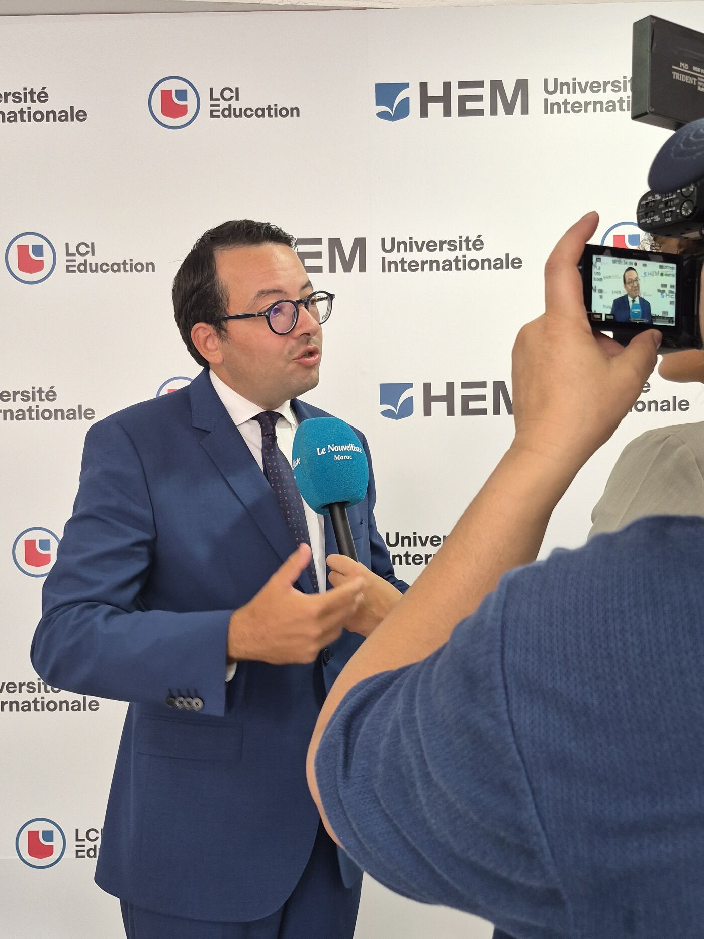 A man in a suit and tie speaks into a microphone while being filmed by a camera, with a backdrop featuring the LCI Education and HEM University logos.