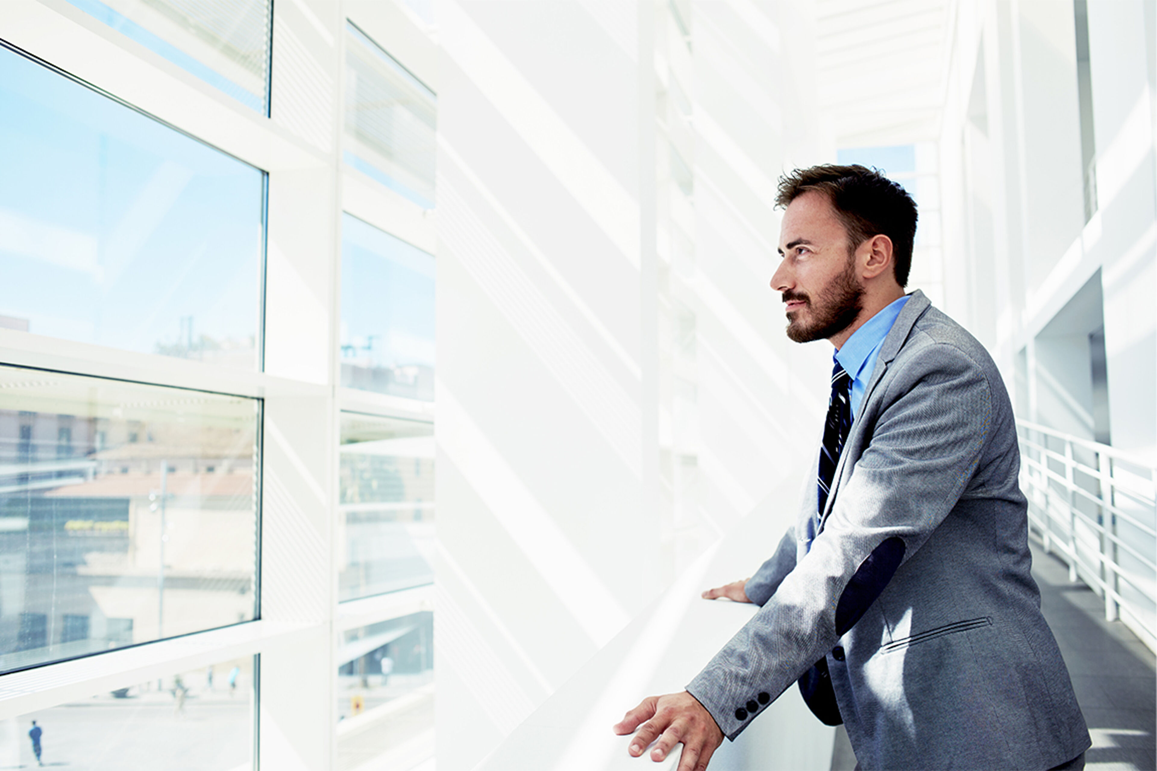 A pensive businessman in a suit looks out a bright office window, overlooking an urban landscape.