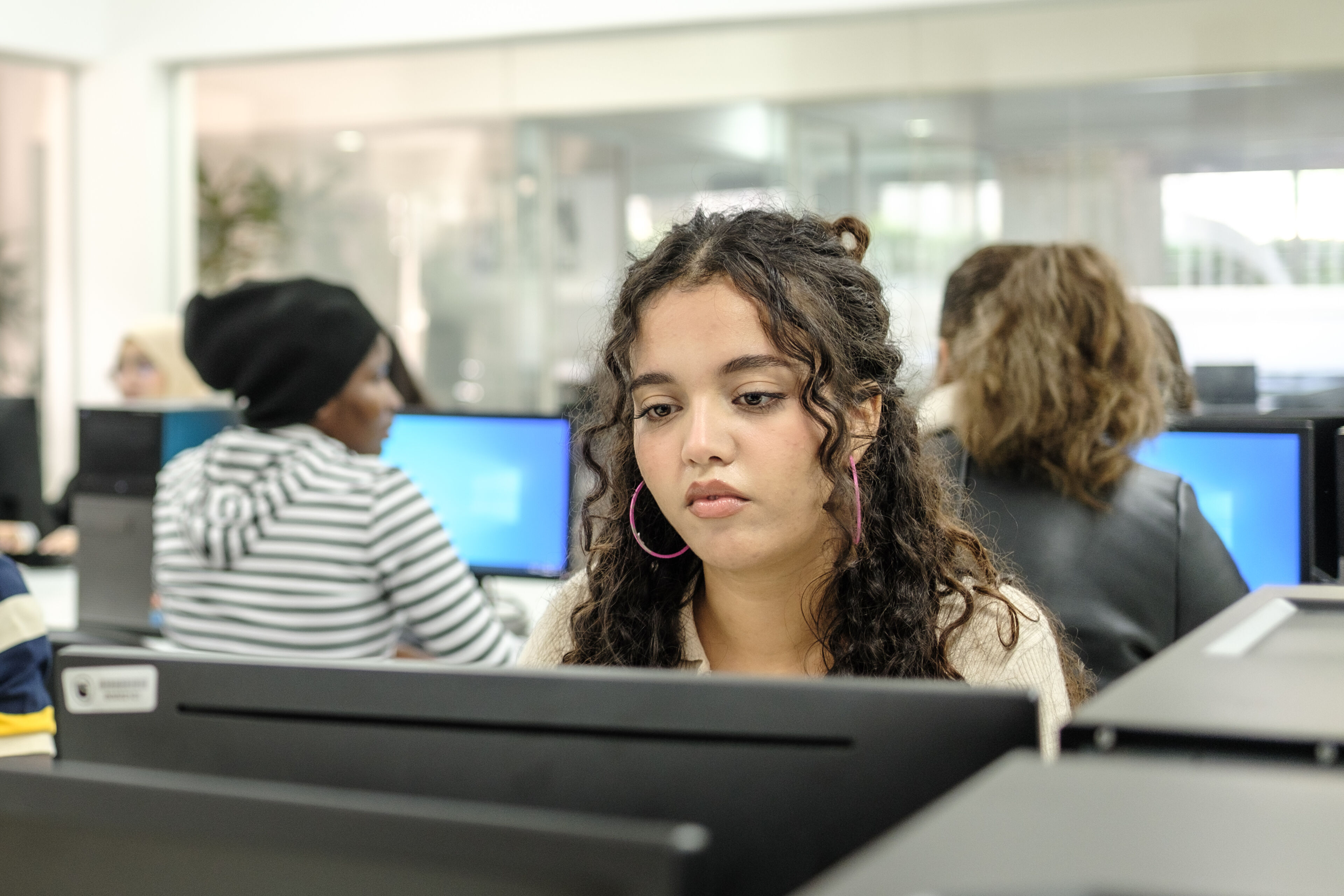 Une jeune femme concentrée aux cheveux bouclés et portant des boucles d'oreilles en forme d'anneau travaille sur un ordinateur dans un laboratoire informatique animé.