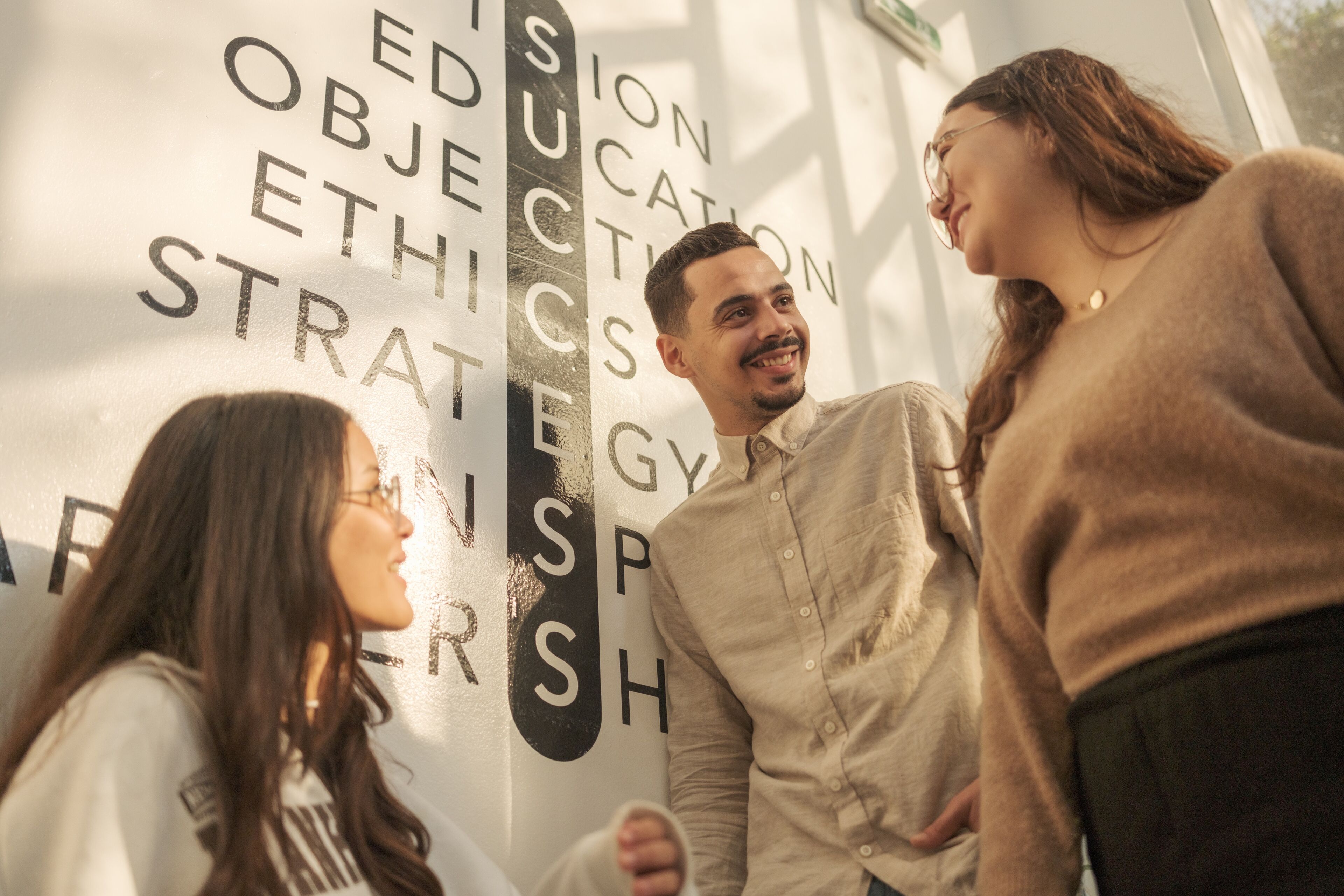 Three students converse joyfully in front of a wall with educational words.