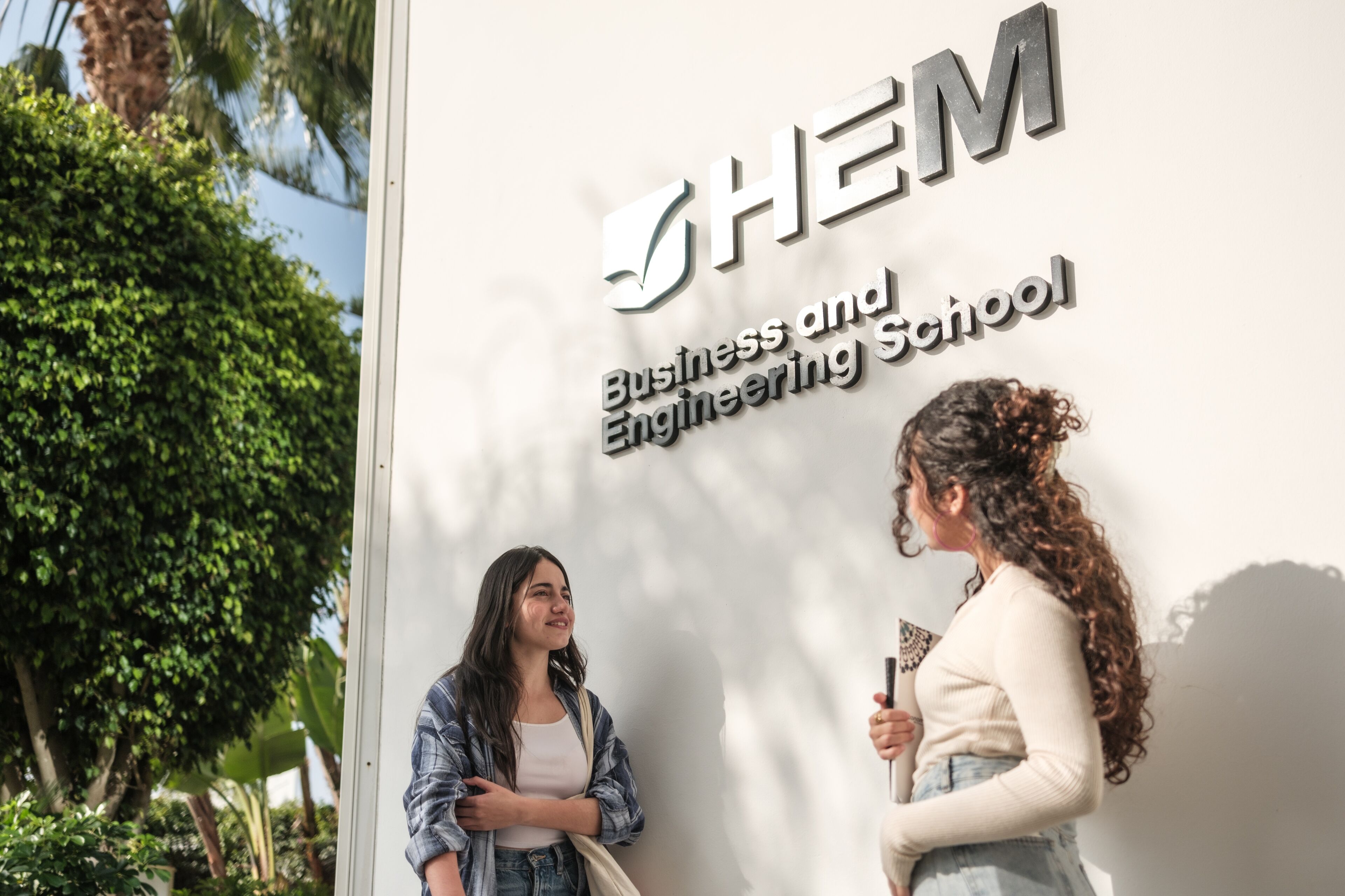 Two students converse by the school sign, "HEM Business and Engineering School," in a sunny outdoor setting with greenery.