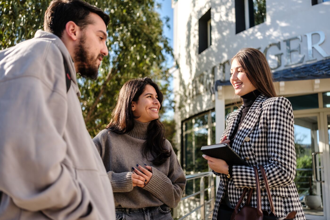 Three college students engage in a friendly conversation on a sunny campus walkway, with one holding a digital tablet.