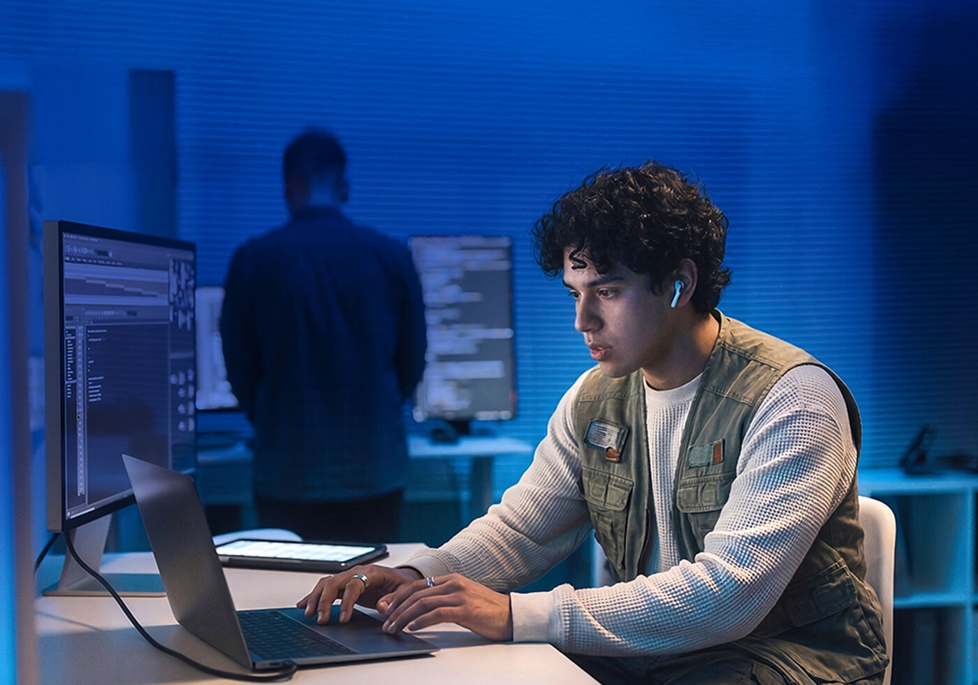 A young man wearing earbuds is focused on his laptop in a dimly lit room with blue lighting. Another person stands in the background.