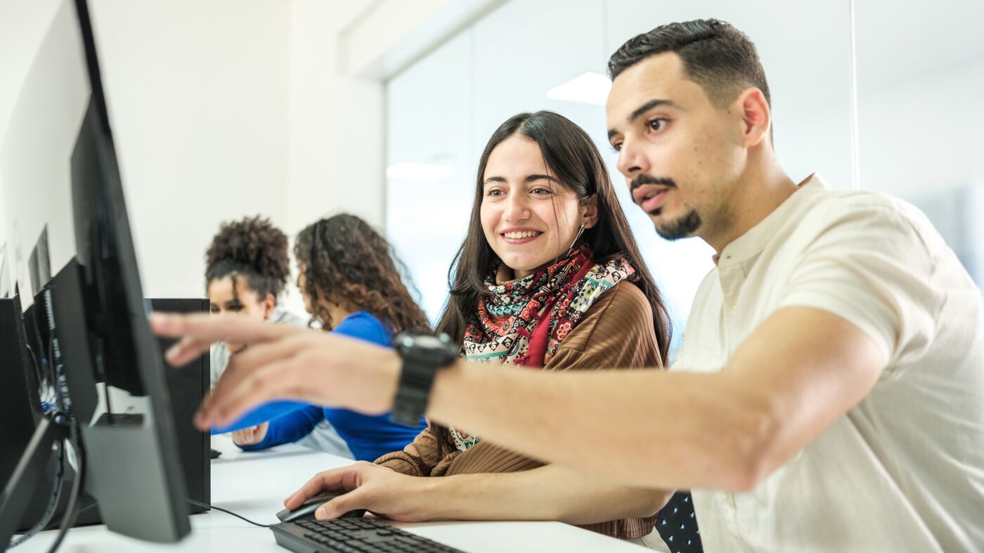 A young man and woman are engaging in a collaborative task at a computer in an office setting, with peers working in the background.