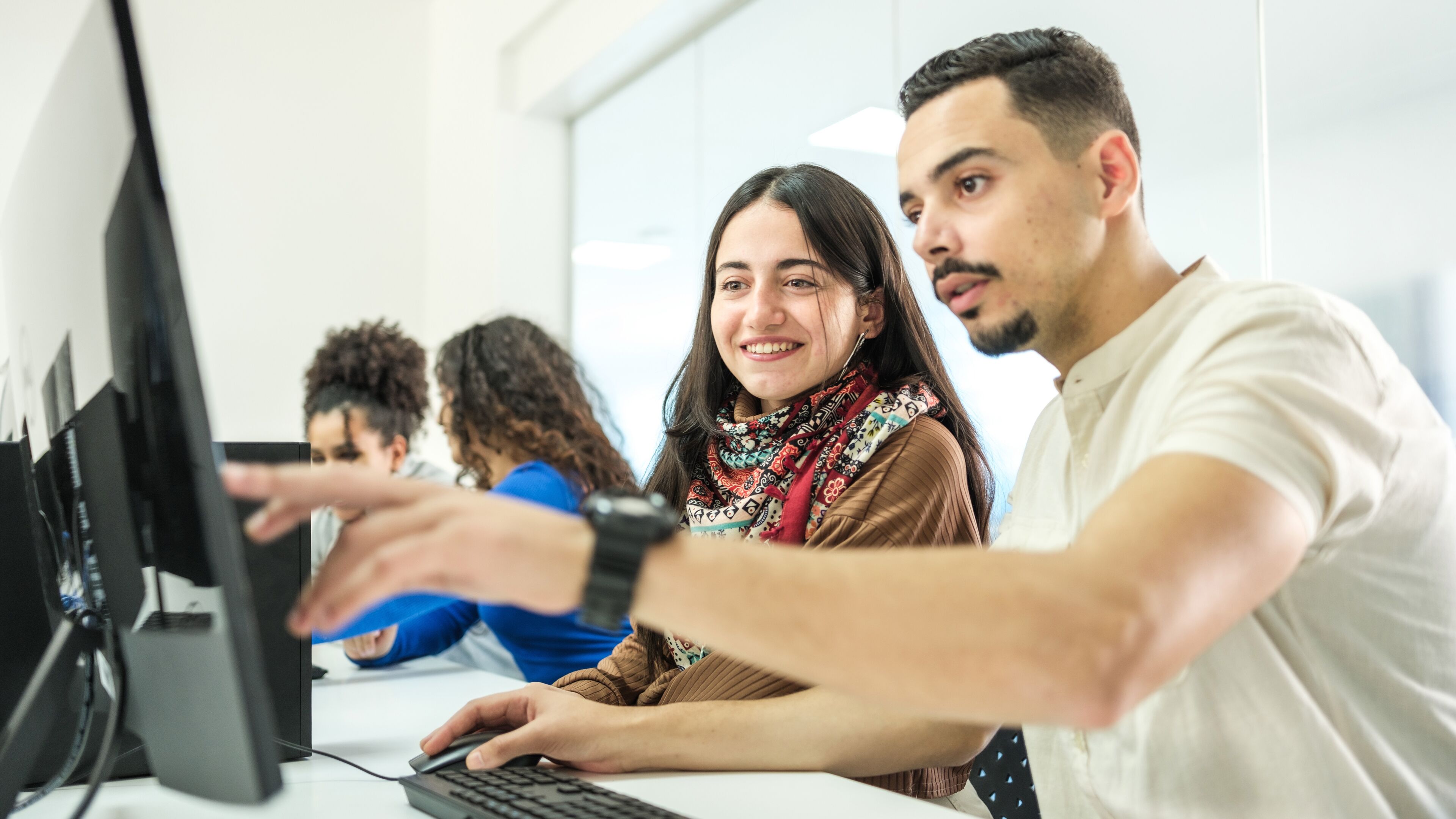 A young man and woman are engaging in a collaborative task at a computer in an office setting, with peers working in the background.