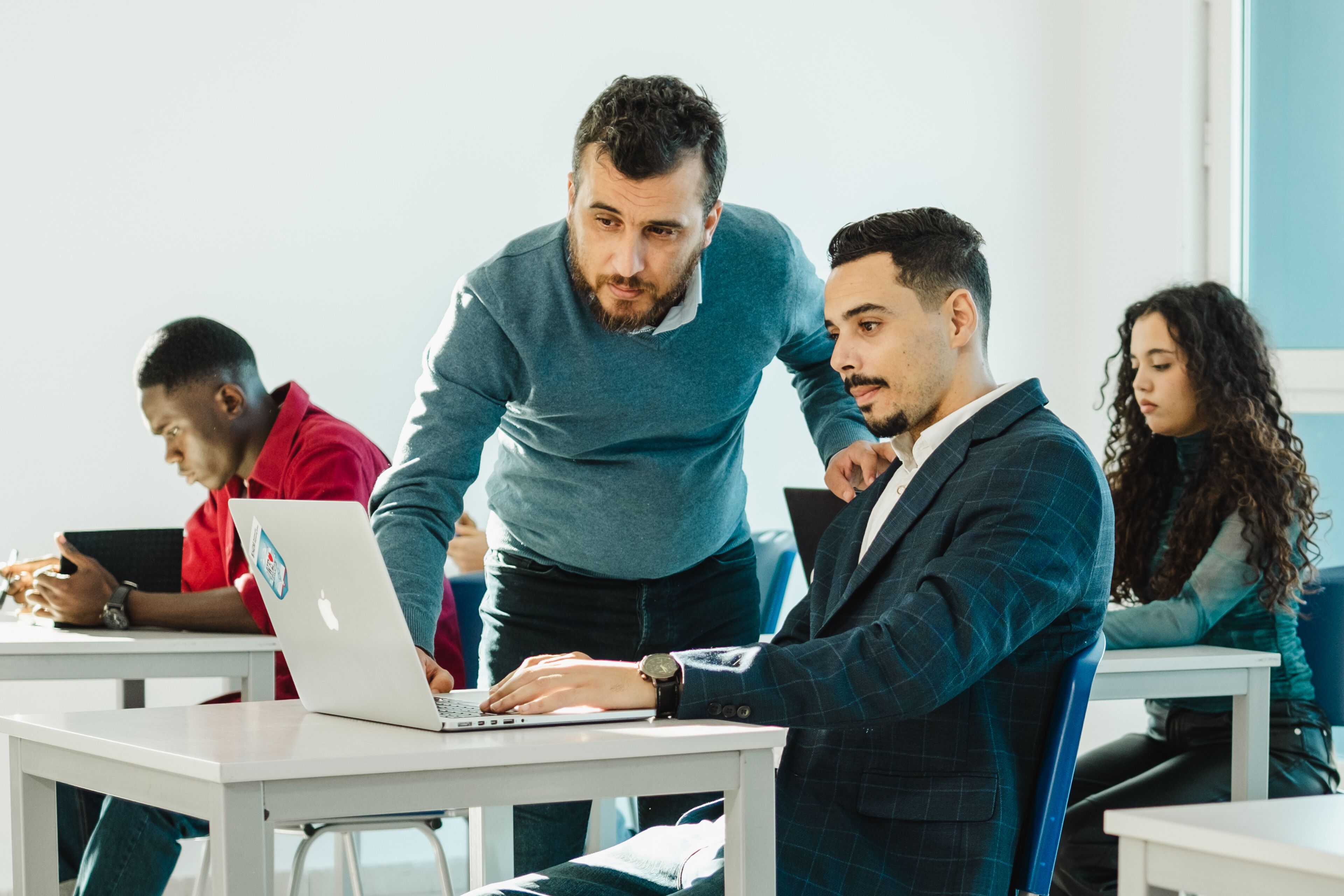 A business instructor guiding an employee on a laptop, with focused colleagues in the background.