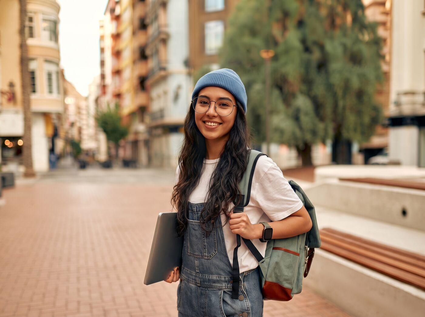A young woman wearing a blue beanie and glasses carries a backpack and a laptop, smiling at the camera on an urban street.