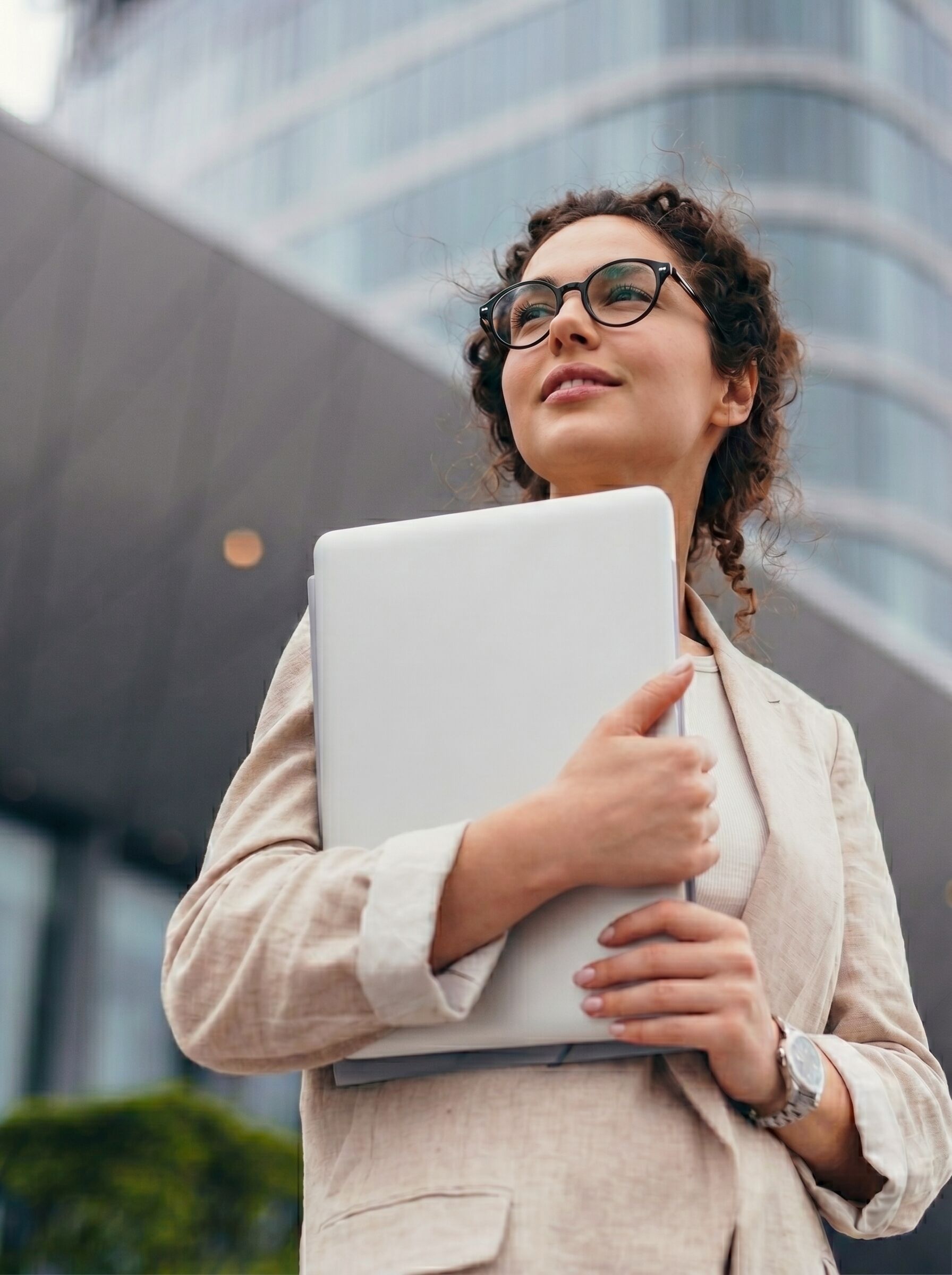 A young woman wearing glasses and a summer suit holds a laptop in front of a modern building, looking upwards with a hopeful expression.