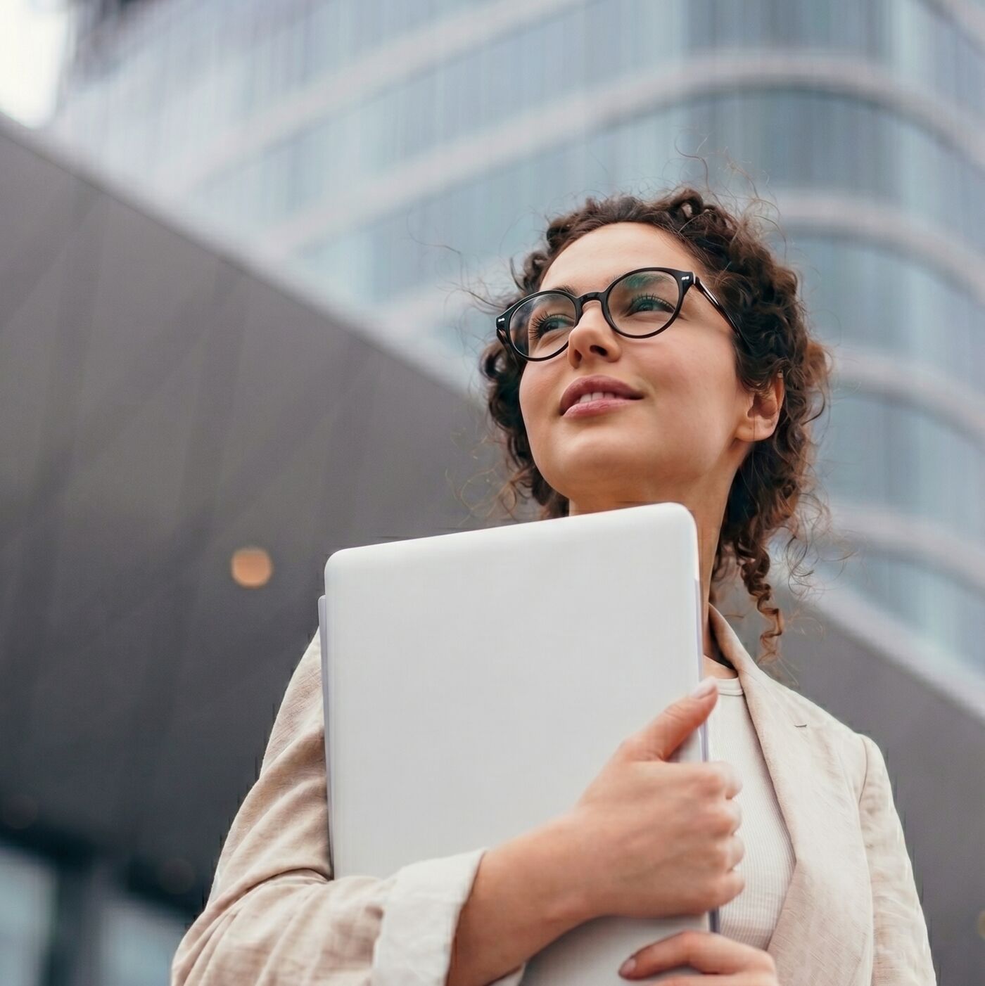 A young woman wearing glasses and a summer suit holds a laptop in front of a modern building, looking upwards with a hopeful expression.