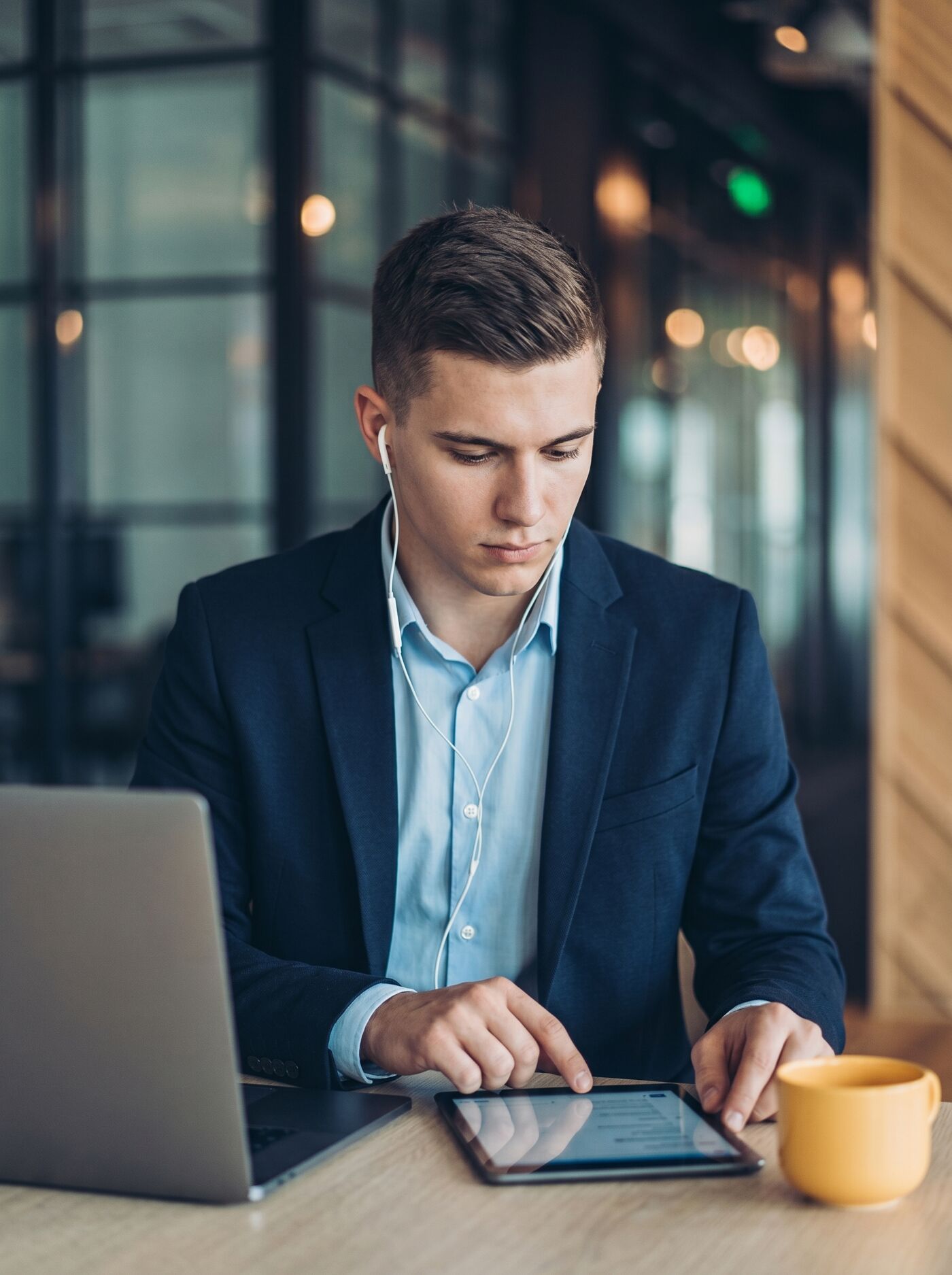 A young businessman wearing earphones is focused on his tablet, with a laptop and a yellow mug on the table.