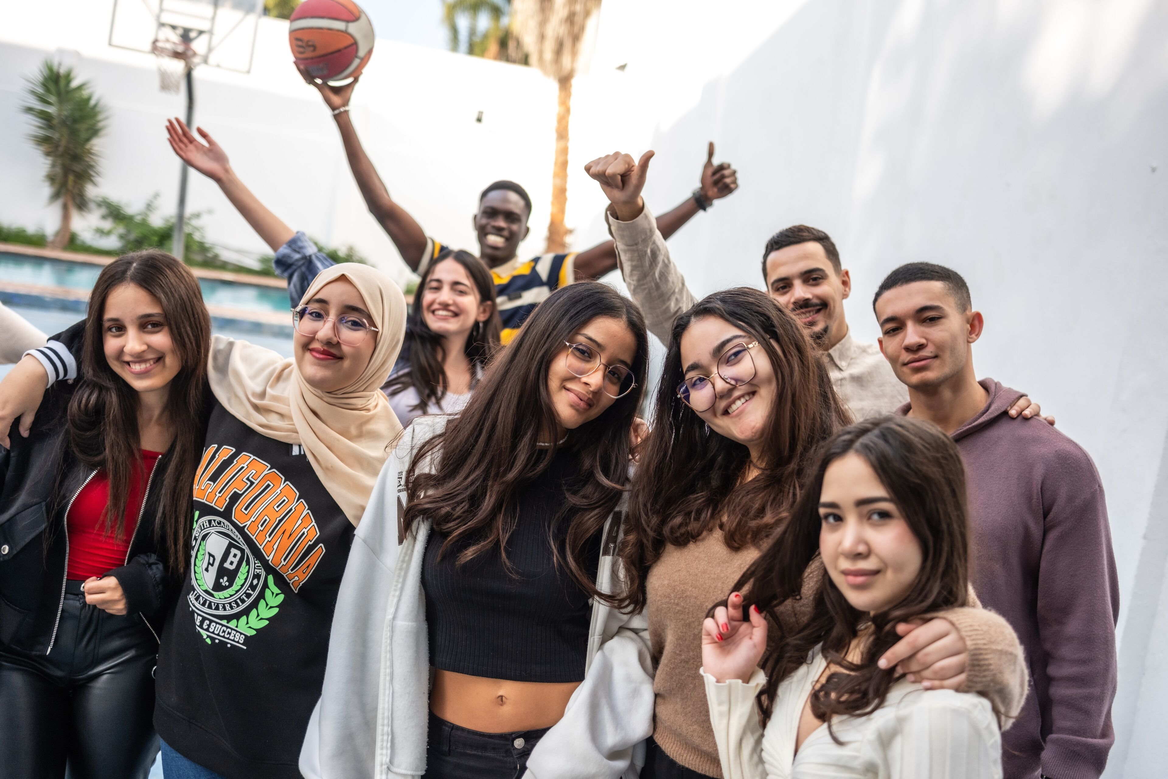 A multiethnic group of young adults share a joyful moment on a sunny day at a basketball court, with some posing and one holding a basketball aloft.