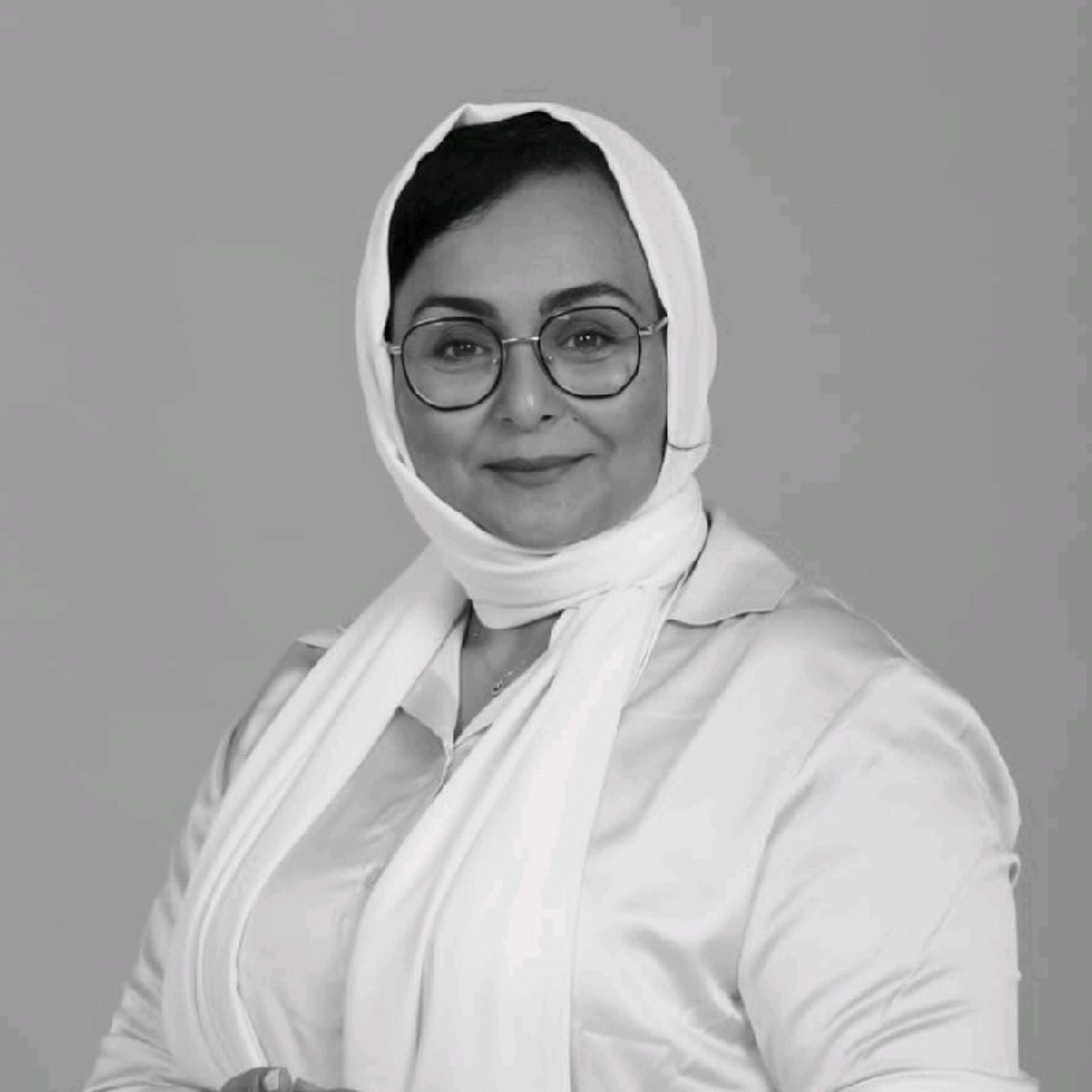 Personne posée avec des lunettes, portant un hijab blanc et un blazer blanc sur un chemisier, avec un sourire doux.