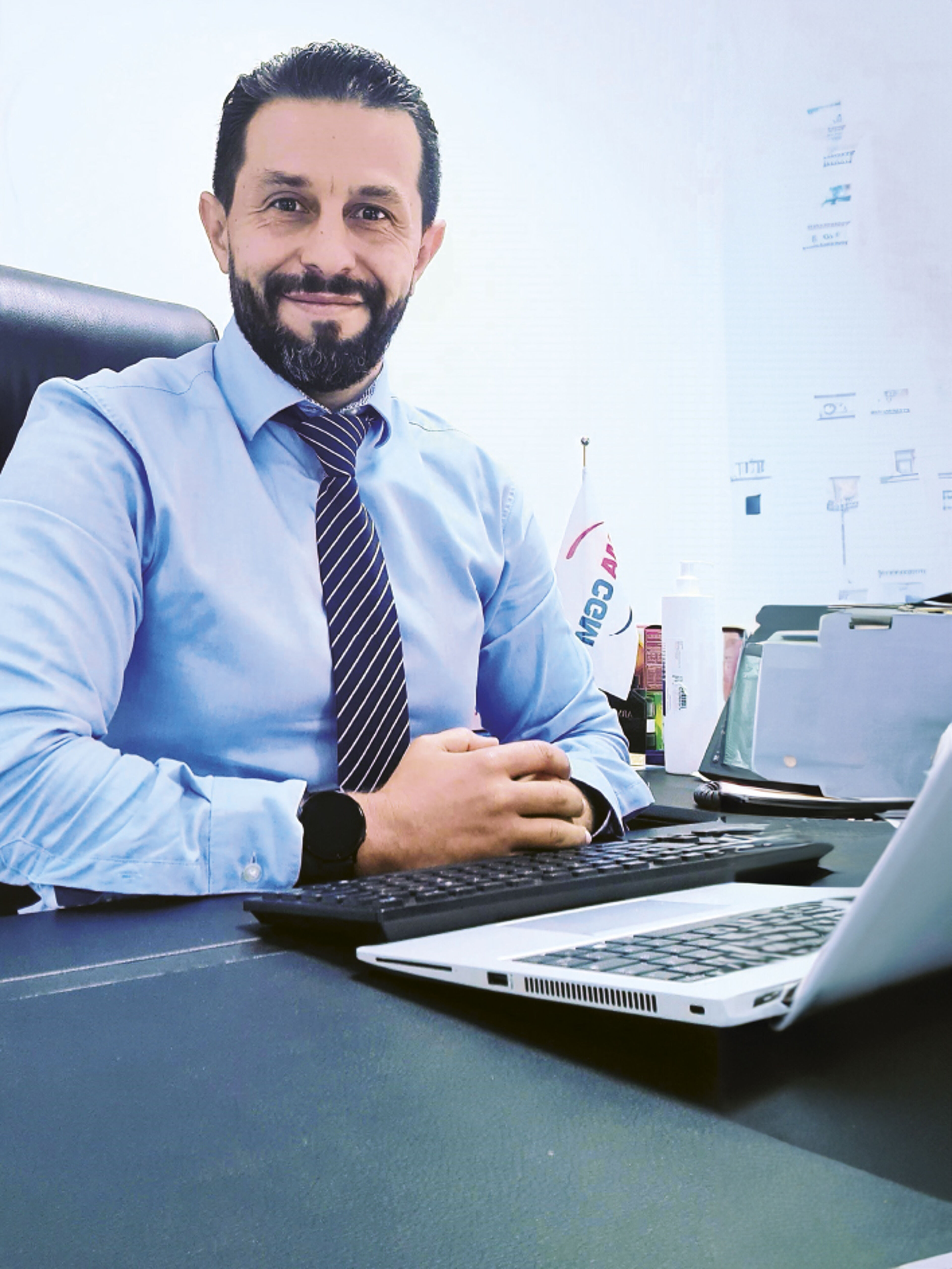Un homme barbu en chemise bleue et cravate rayée sourit assis à son bureau avec un ordinateur portable, dégageant confiance et accessibilité.