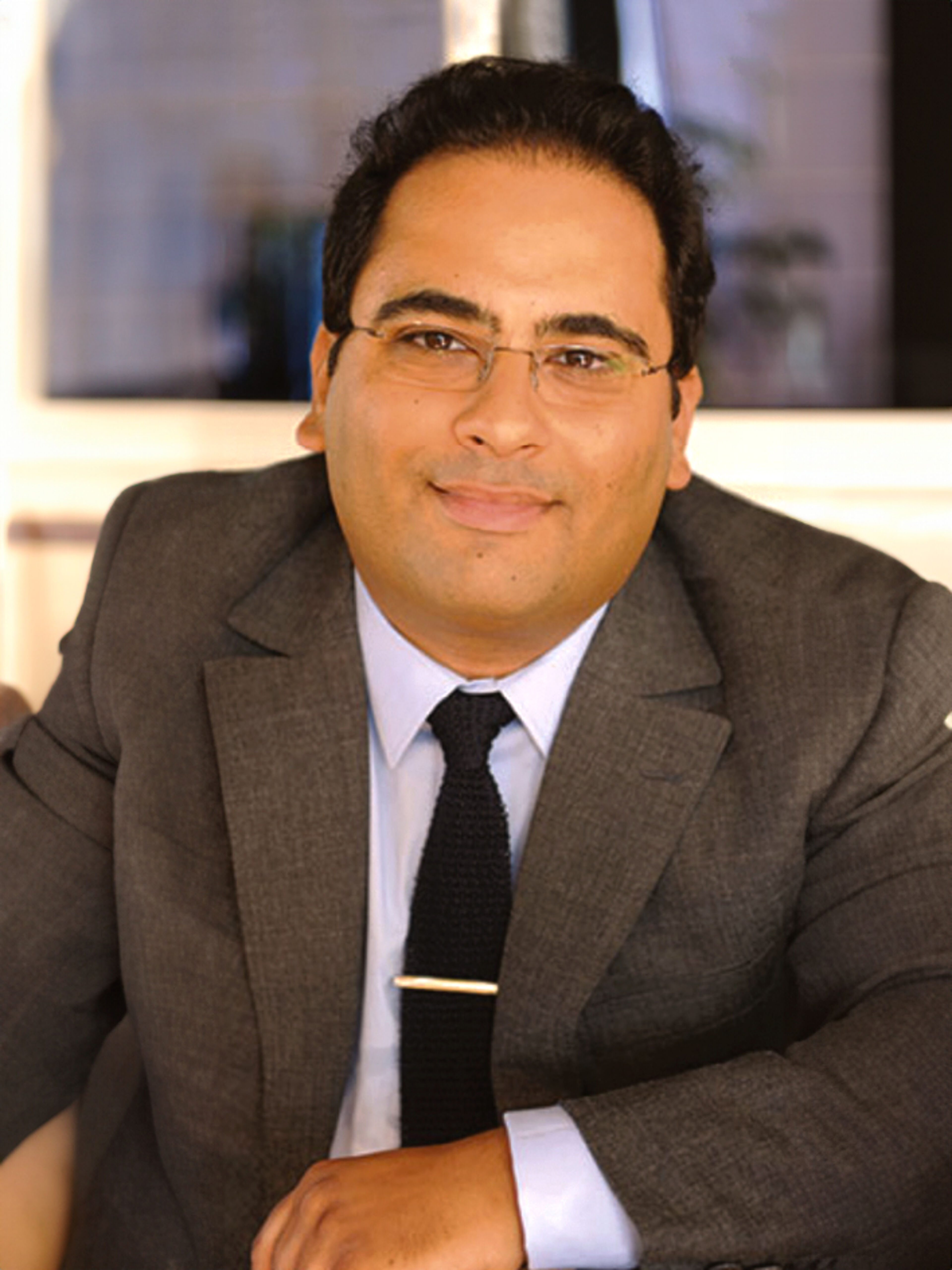 Professionnel masculin confiant en costume gris, chemise blanche et cravate noire, portant des lunettes, sur fond intérieur flou.