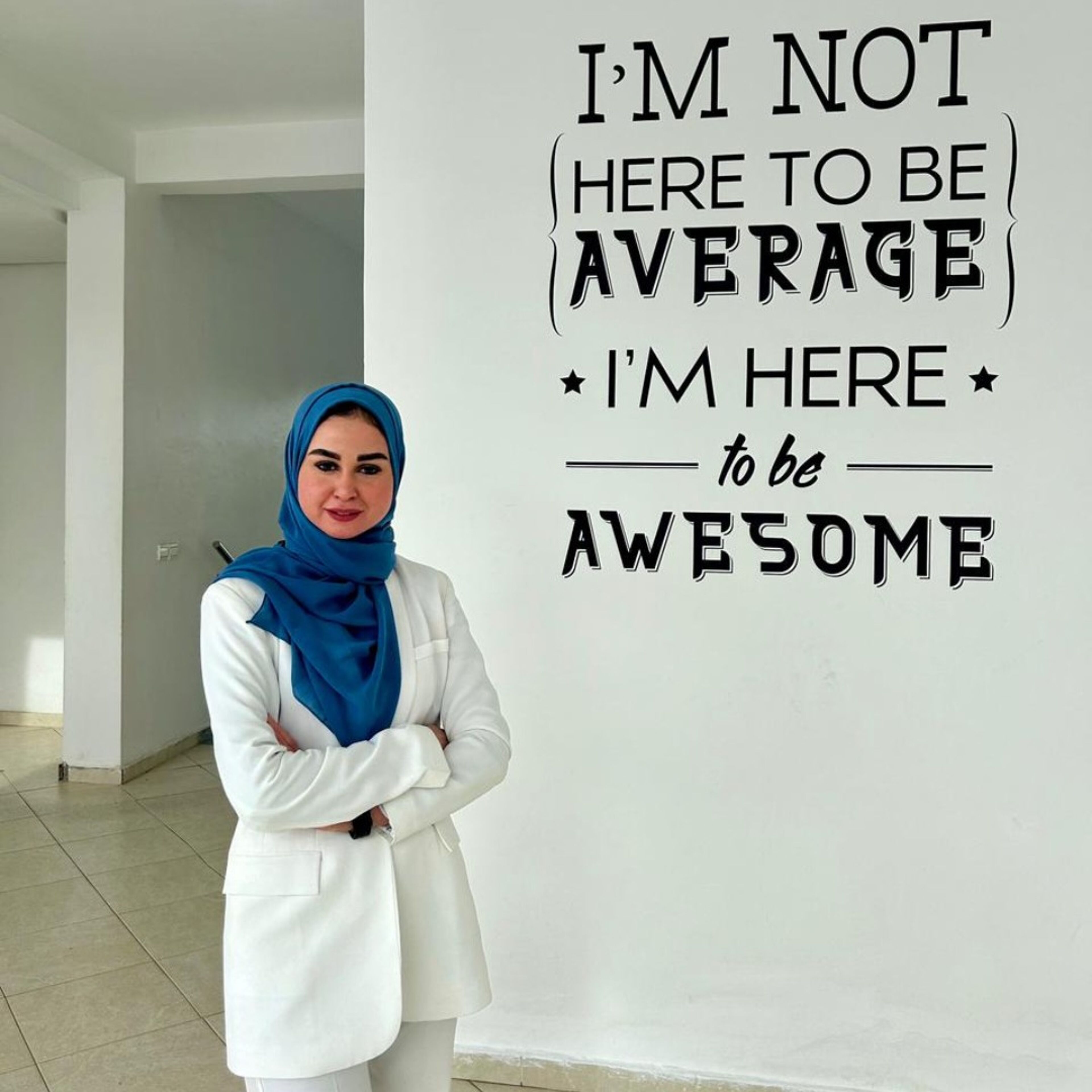 Une femme posée en manteau blanc et hijab bleu se tient fièrement devant un mur portant la citation motivante "JE NE SUIS PAS ICI POUR ÊTRE MOYENNE, JE SUIS LÀ pour être GÉNIALE".