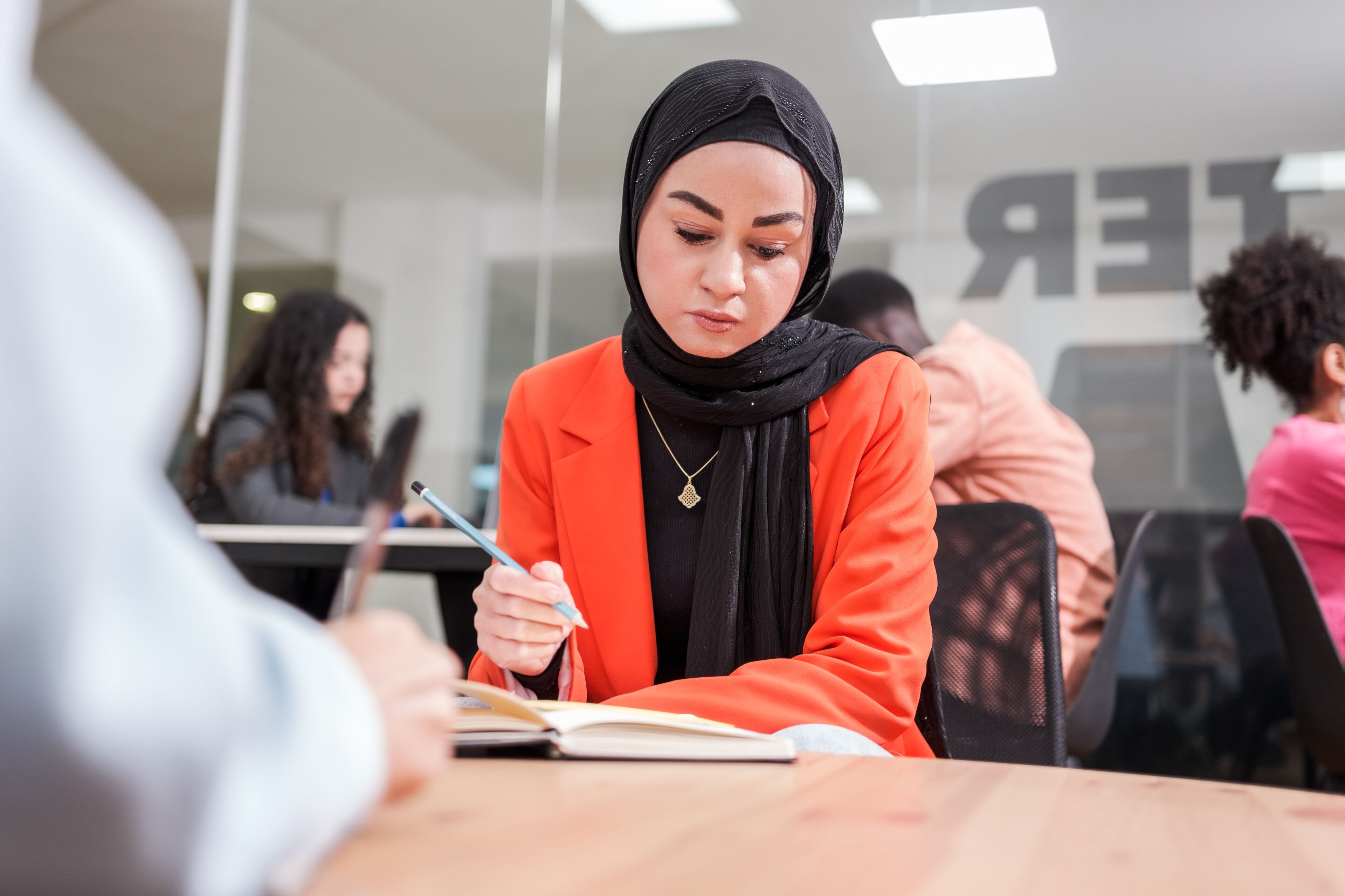 Une femme concentrée en hijab prenant des notes à une table d'étude animée.