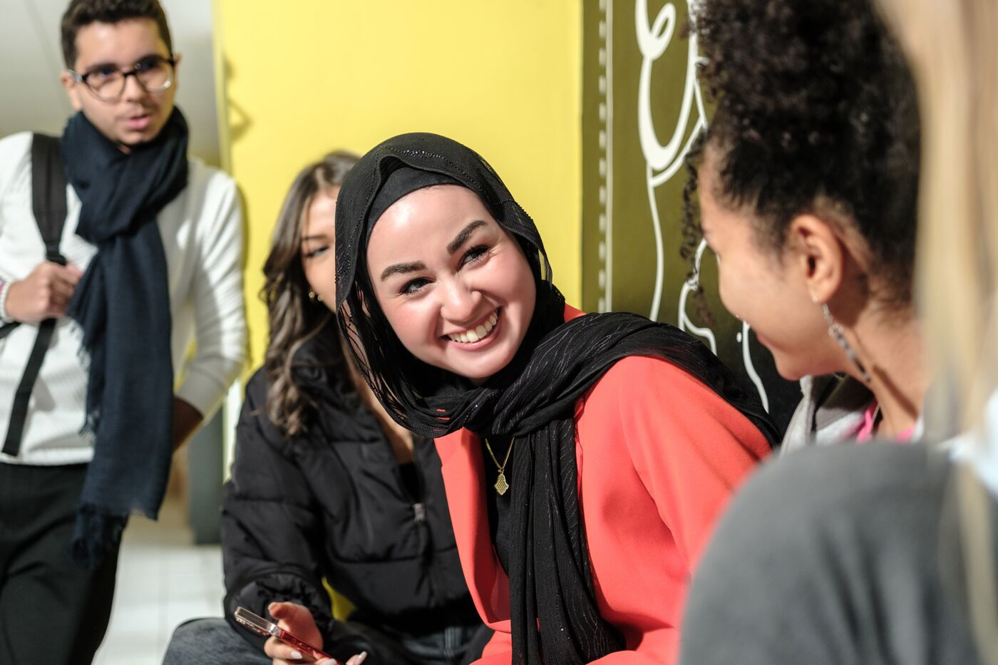 A cheerful young woman in a black hijab and red top enjoys a moment with her multicultural peers in a casual setting.