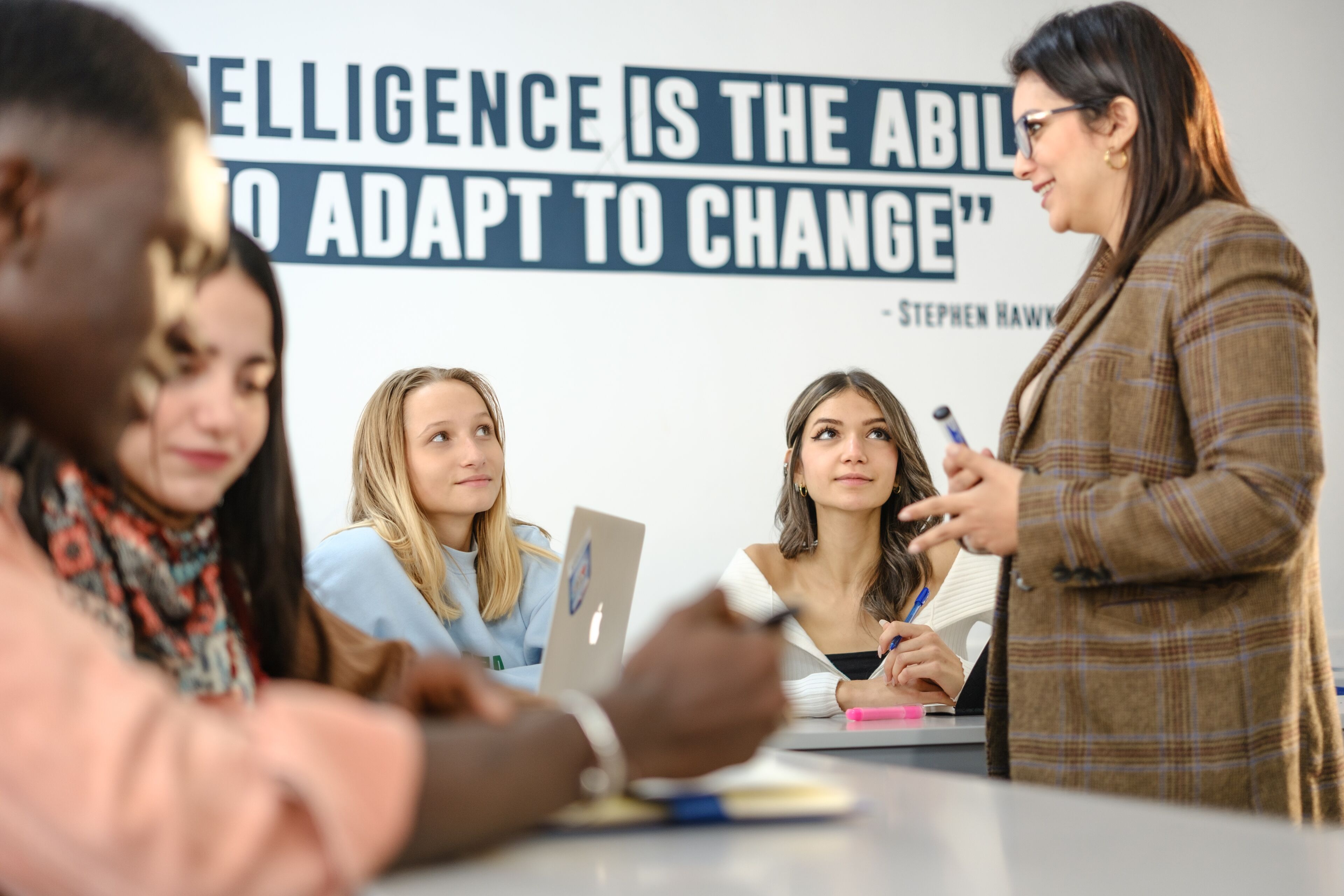 A diverse group of students attentively listening to a teacher in a classroom, with a quote by Stephen Hawking on the wall about adaptability and intelligence.