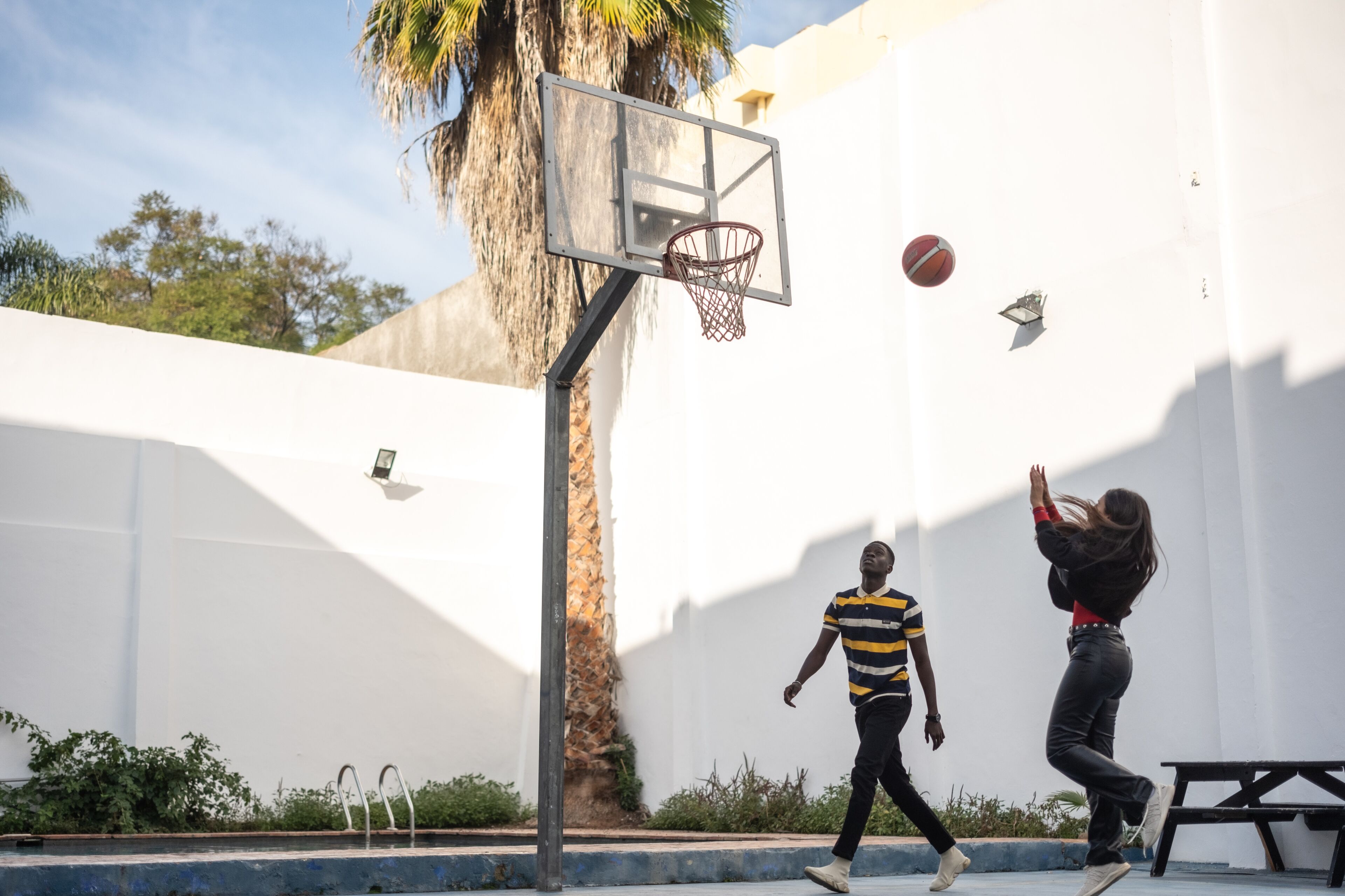 Deux personnes jouent au basketball dans un terrain extérieur, l'une tire le ballon vers le panier.